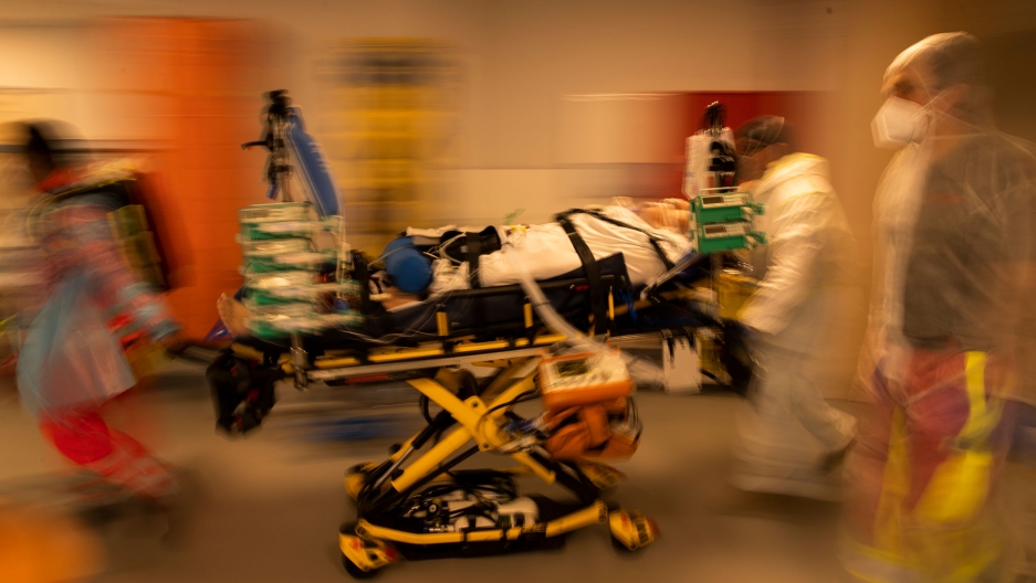 Medical personnel wheel a bed with a coronavirus patient in critical condition as they prepare to transfer the patient by ambulance to a hospital in Aachen, Germany, April 14, 2021. A COVID-19 patient is shown strapped to a yellow gurney with several medical staff moving along side in a blurred motion photograph.