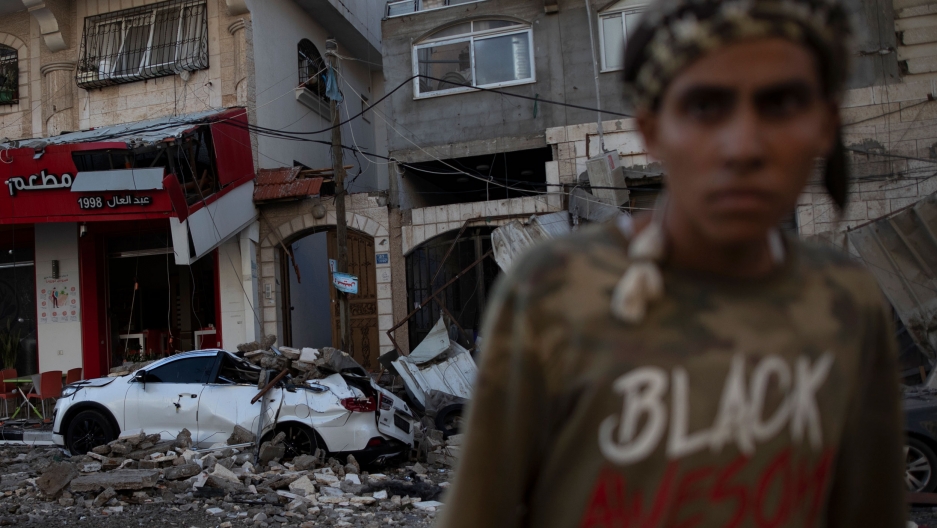 Damaged building are seen following Israeli airstrikes on Gaza City, May 12, 2021. A person is shown in soft focus wearing a shirt with the word "Black" printed on it with the rubble of a bombed building a car in the background.