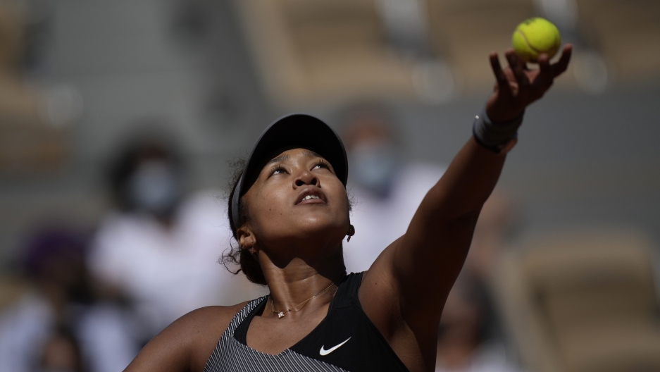 Japan's Naomi Osaka serves the ball to Romania's Patricia Maria Tig during their first round match of the French Open tennis tournament at the Roland Garros stadium Sunday, May 30, 2021, in Paris. A tennis player lifts her ball and racket into the air.