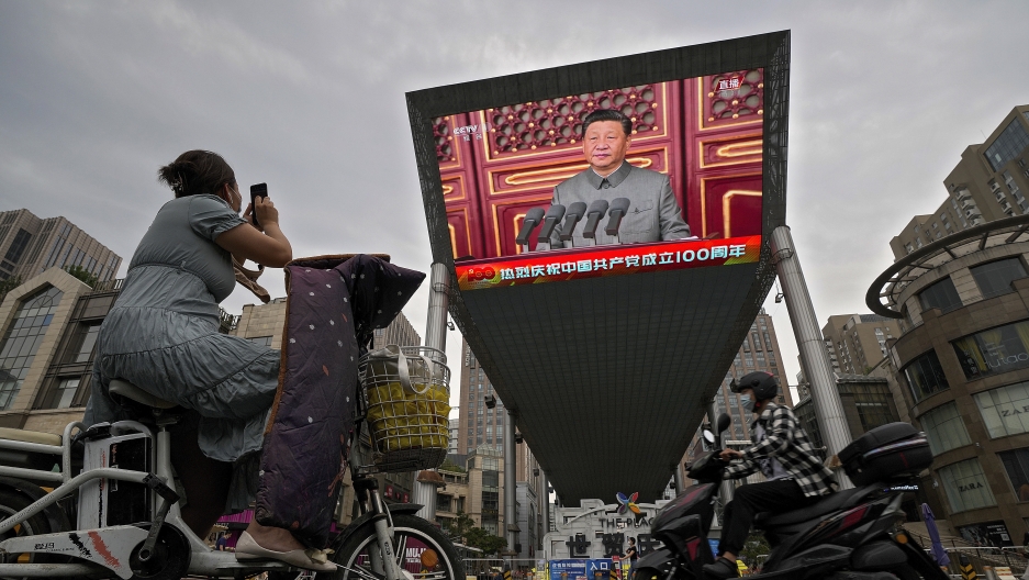 A woman on her electric-powered scooter films a large video screen outside a shopping mall showing Chinese President Xi Jinping speaking during an event to commemorate the 100th anniversary of China's Communist Party at Tiananmen Square in Beijing, Thursd A woman on her electric-powered scooter films a large video screen outside a shopping mall showing Chinese President Xi Jinping speaking during an event to commemorate the 100th anniversary of China's Communist Party at Tiananmen Square in Beijing.