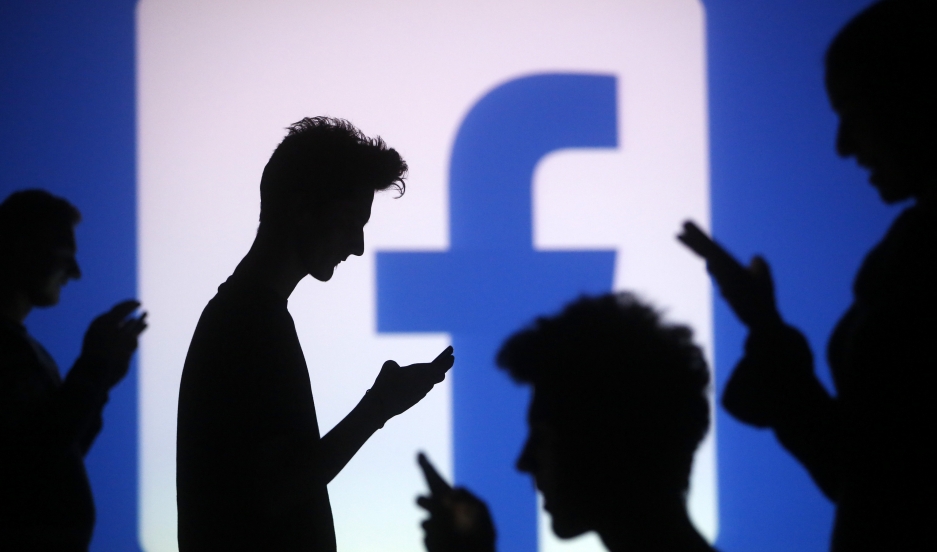 People are silhouetted as they pose with mobile devices in front of a screen projected with a Facebook logo