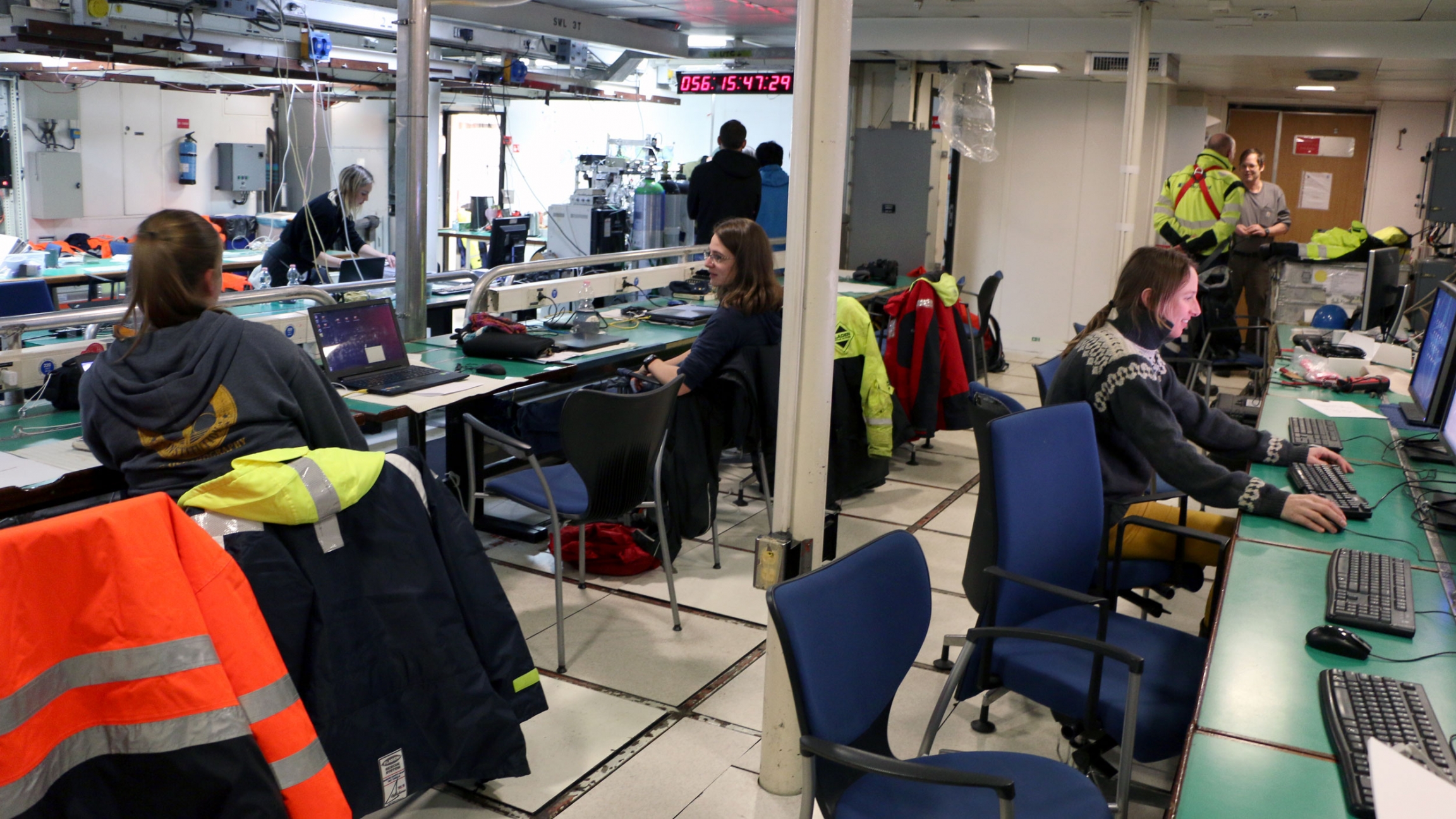 People sit at large tables with computers on the deck of a ship.