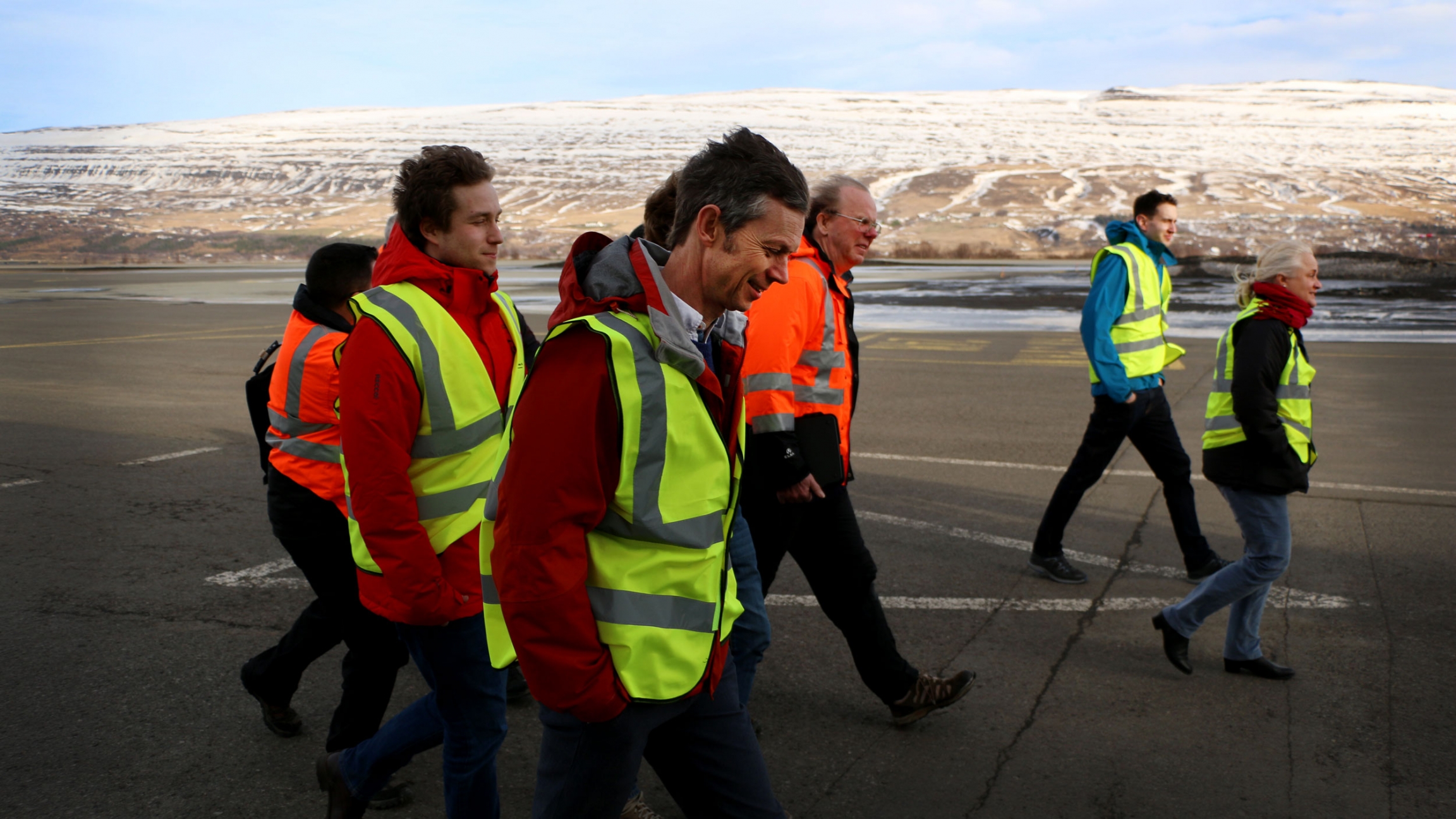 A group of people in bright safety vests walk through the wind on a tarmac.