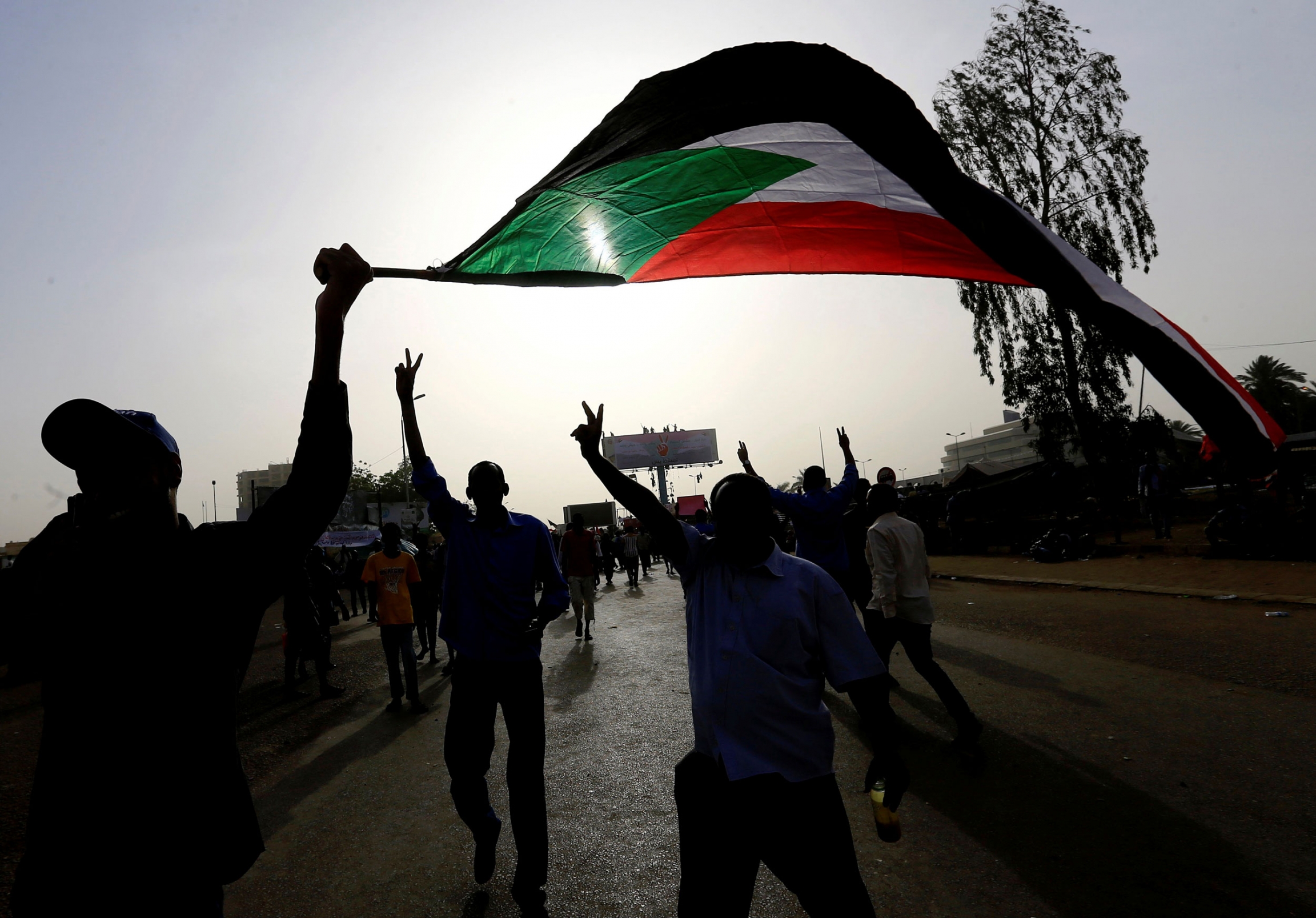 Sudanese demonstrators wave their national flag as they arrive for a protest rally demanding Sudanese President Omar Al-Bashir to step down outside the Defence Ministry in Khartoum, Sudan, on April 11, 2019. People hold their hands in the air and wave a large Sudanese flag. They are backlit by the sun.