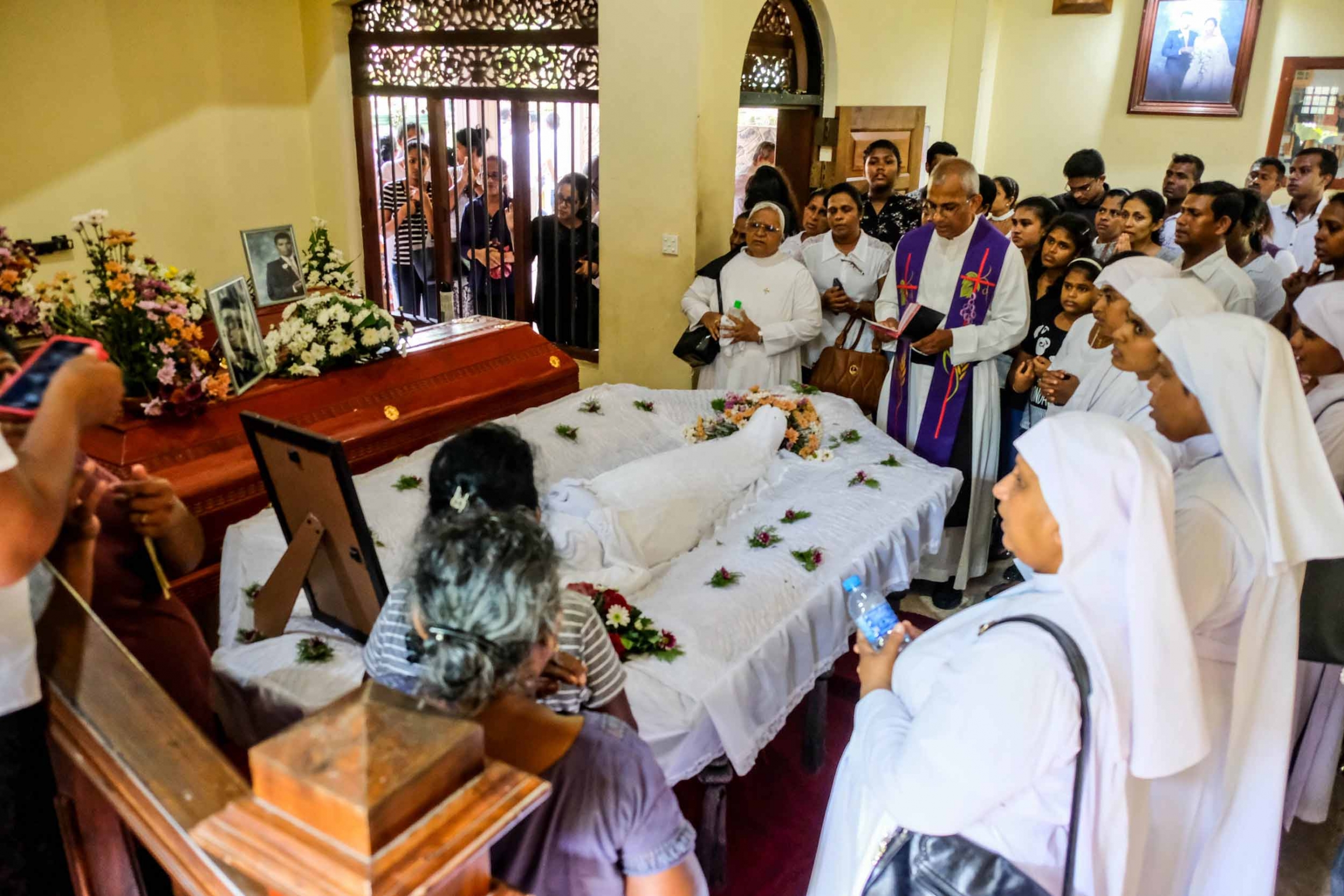 Family and friends gather around a casket in a small home while a priest reads a service
