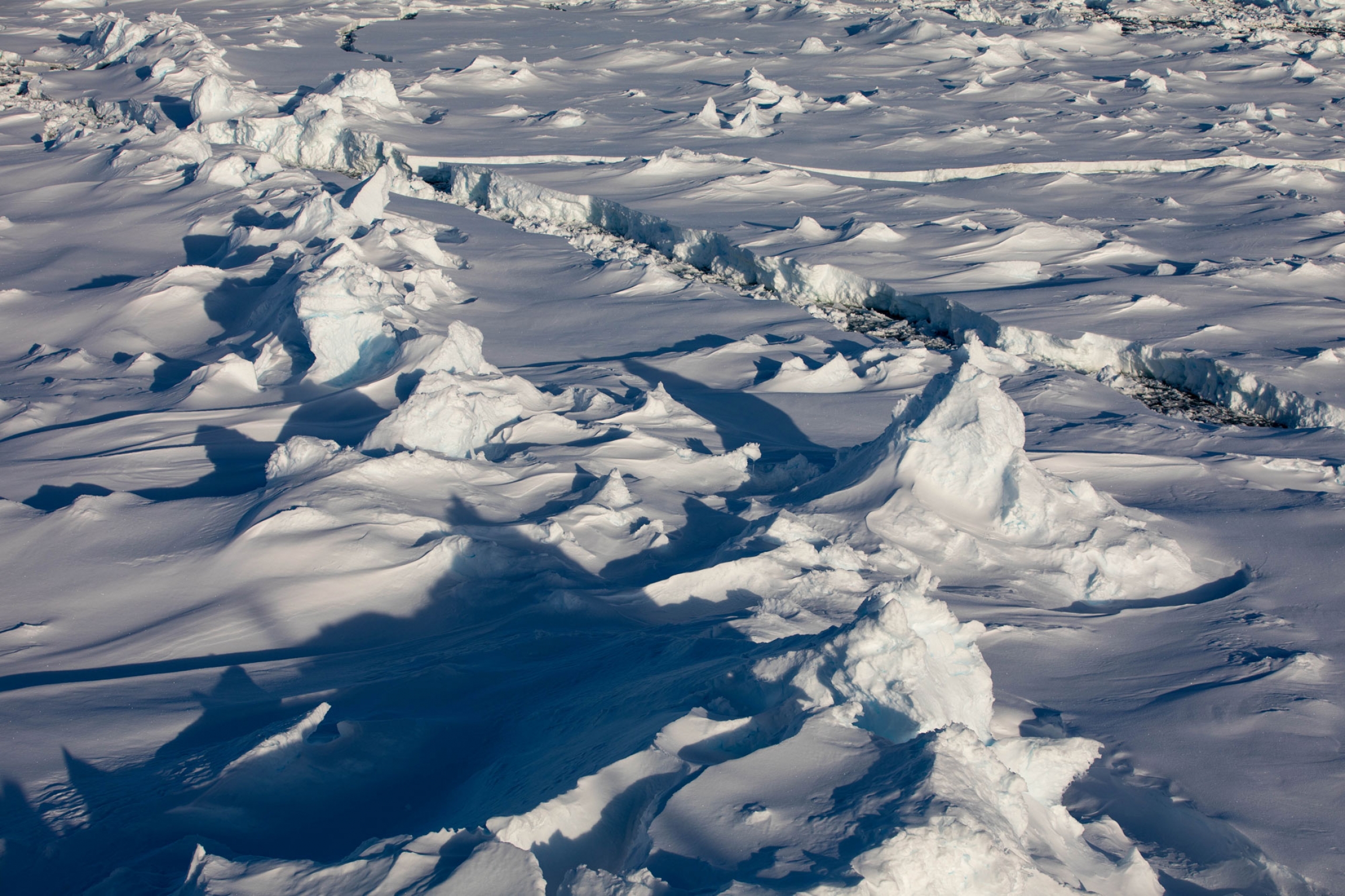 As it ages, pieces of ice crash into each other to form pressure ridges. As it ages, large white pieces of ice crash into each other to form pressure ridges in a photo with a thin water path through the two sides.