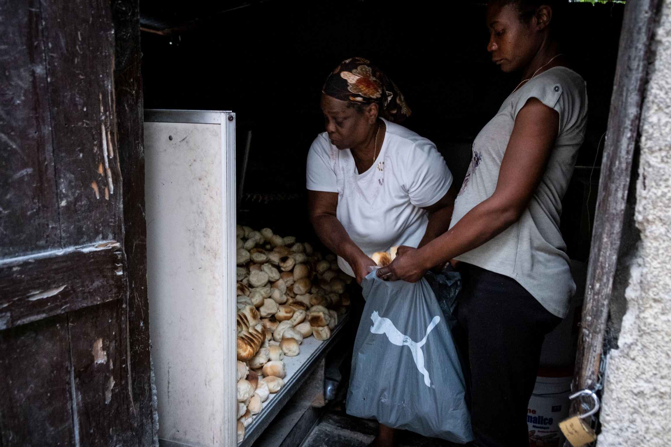 Marie serves a customer inside the bakery next to her home. A woman is shown leaning over helping another woman load with biscuits into a plastic bag.