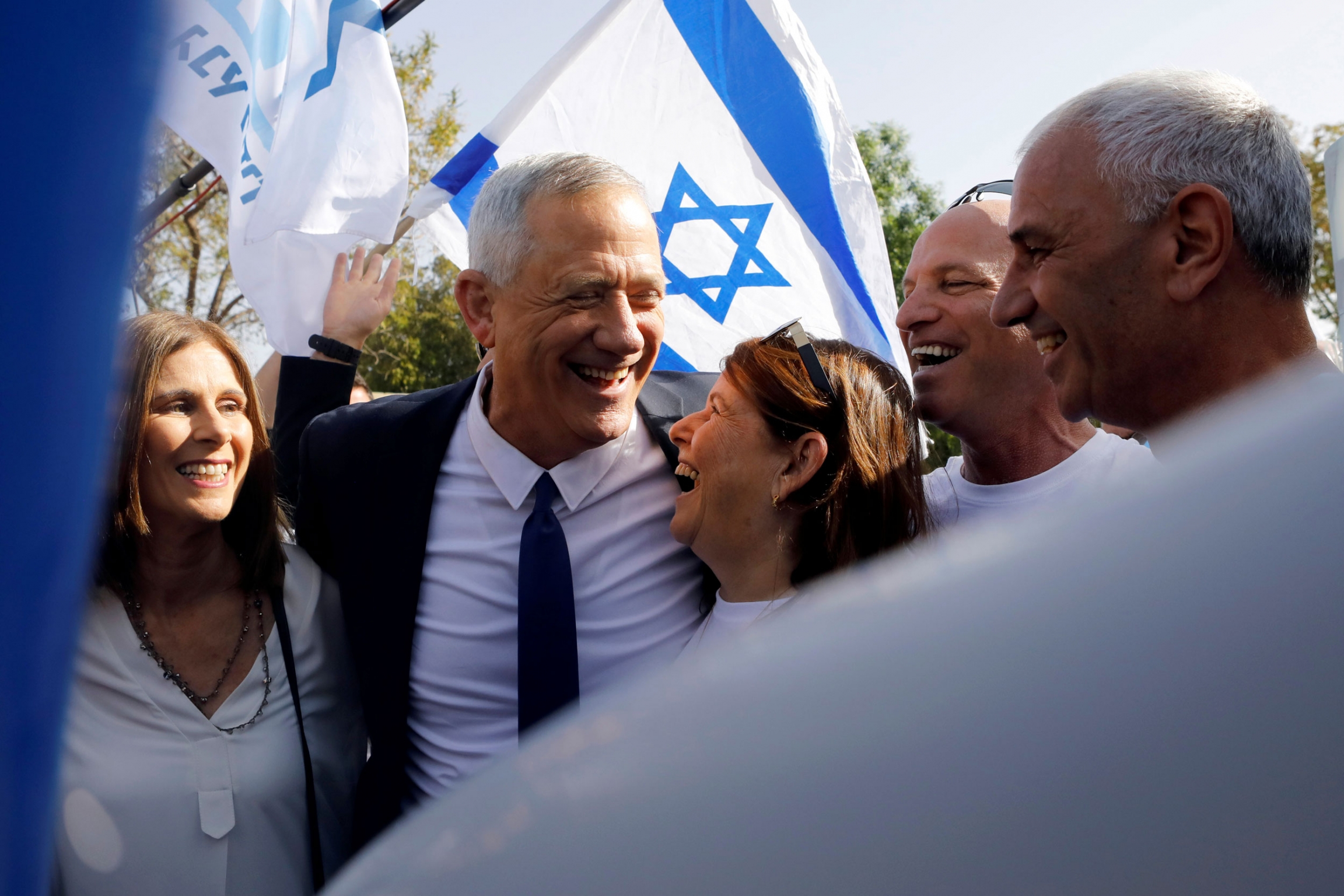 Benny Gantz, leader of Blue and White party, celebrates with supporters and his wife Revital after casting his ballot. Benny Gantz is shown hugging his wife under his arm and an Israeli flag held behind him in the background.