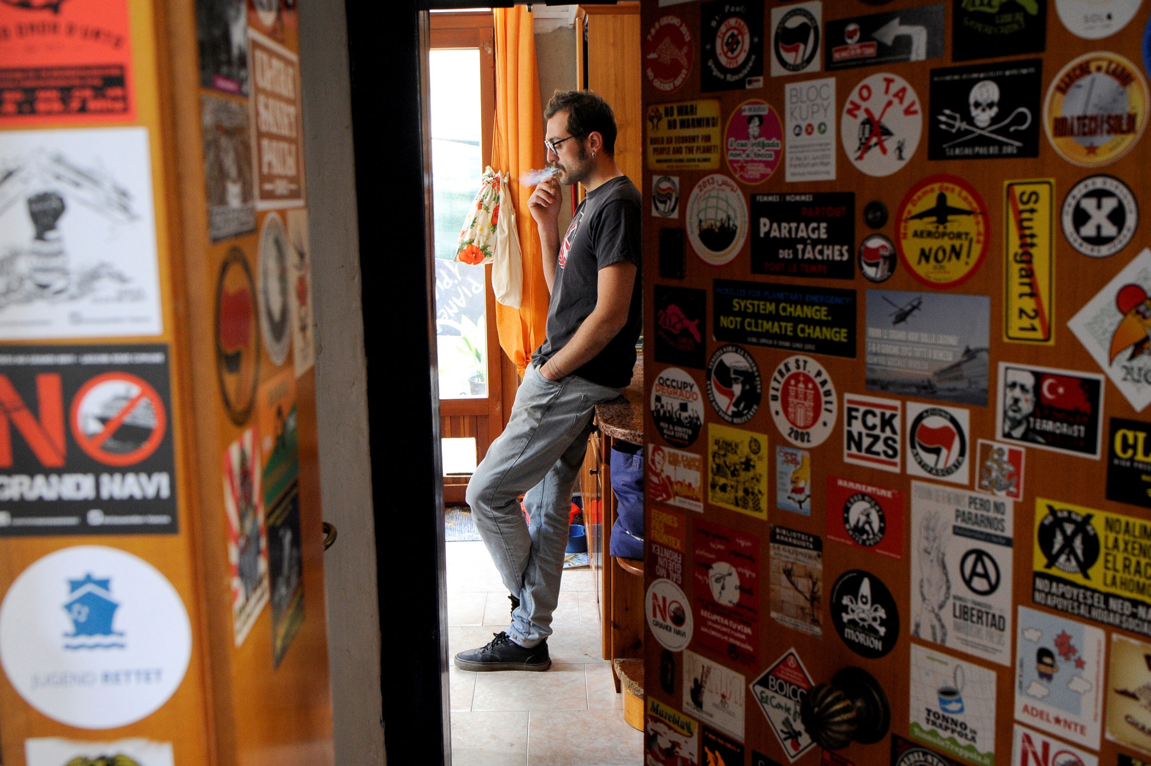Pasquale Ambrogio, 33, smokes a cigarette in his illegally occupied apartment in Venice. A man is seen through a sticker-covered doorway, leaning up against a dresser and smoking a cigarette.