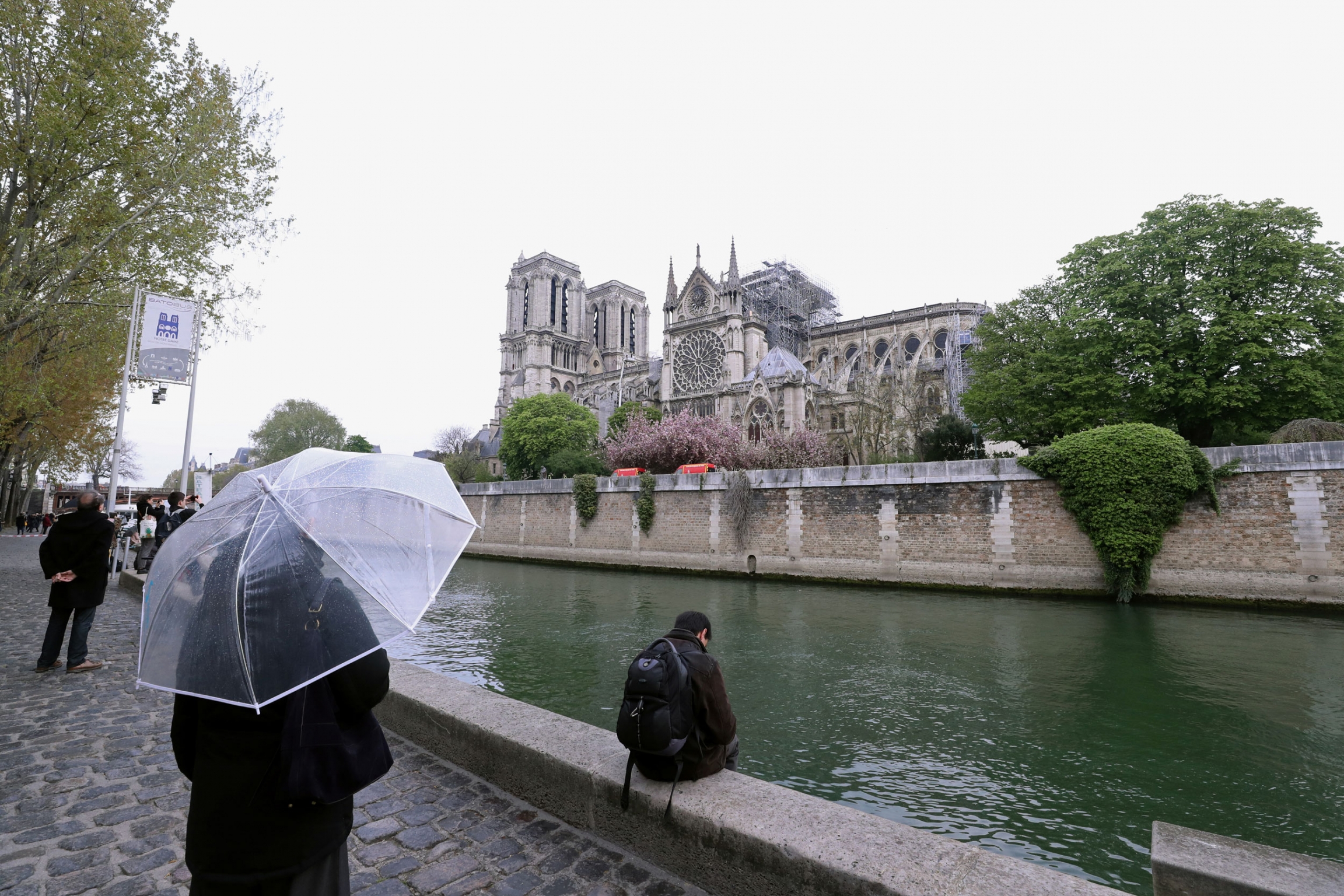People look at Notre-Dame Cathedral after a fire devastated large parts of the gothic gem in Paris, France April 16, 2019. A person holding a clear umbrella stands across the river looking at a burned Notre-Dame Cathedral.