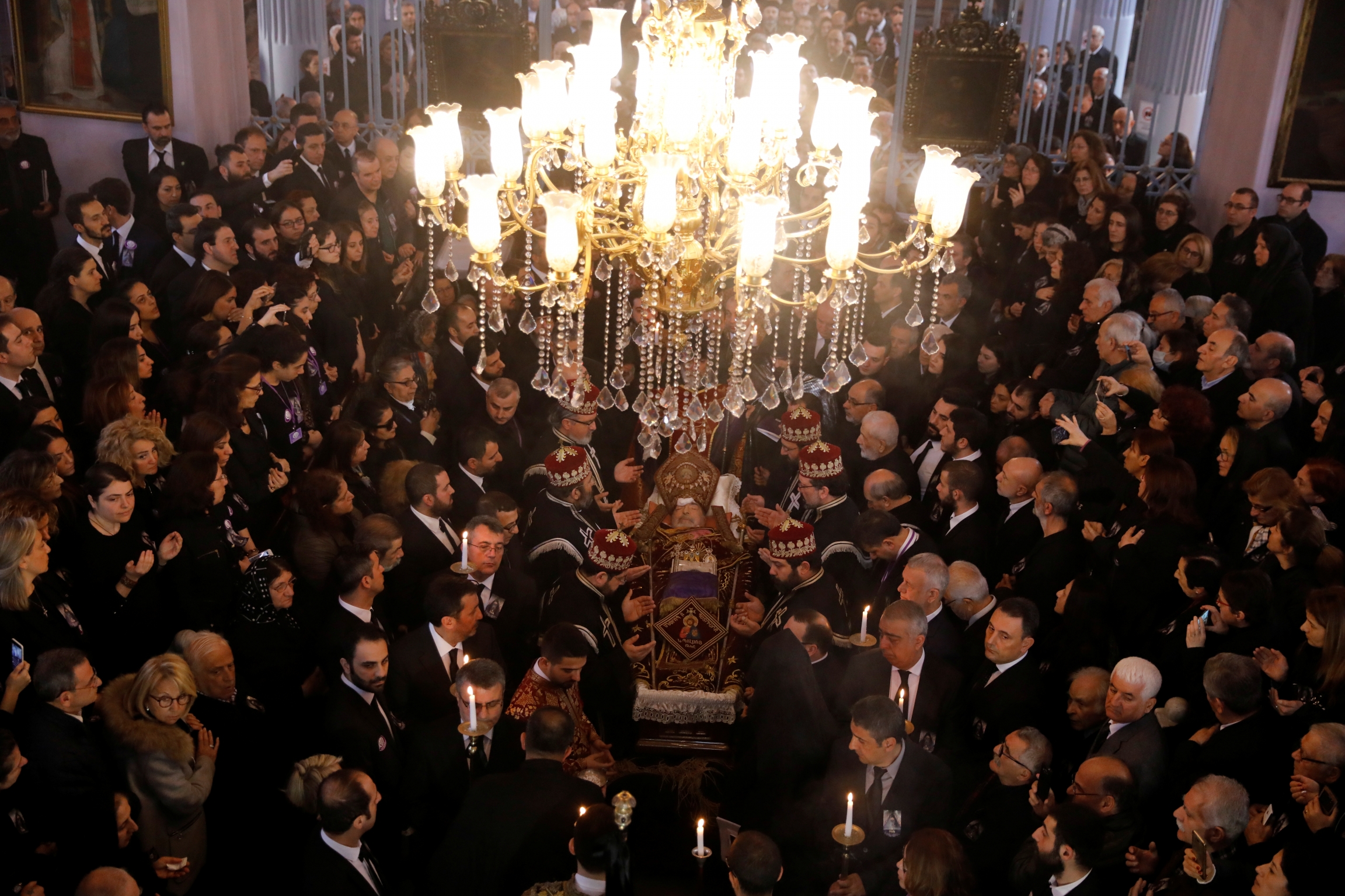 Mourners in black for Armenian Patriach stand under huge chandelier in church.