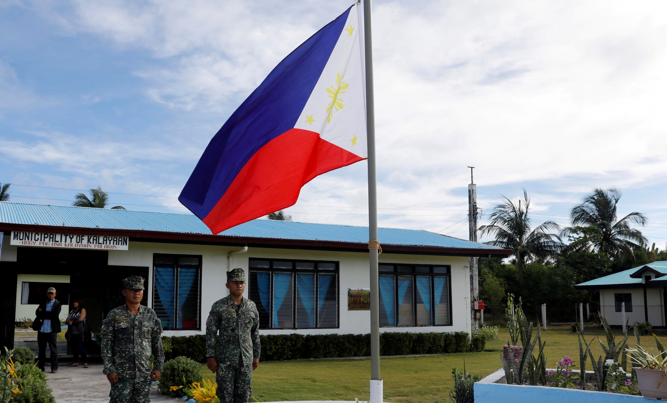 Soldiers stand guard on disputed Thitu island wearing green uniforms and blue, red, white and yellow flag waving.