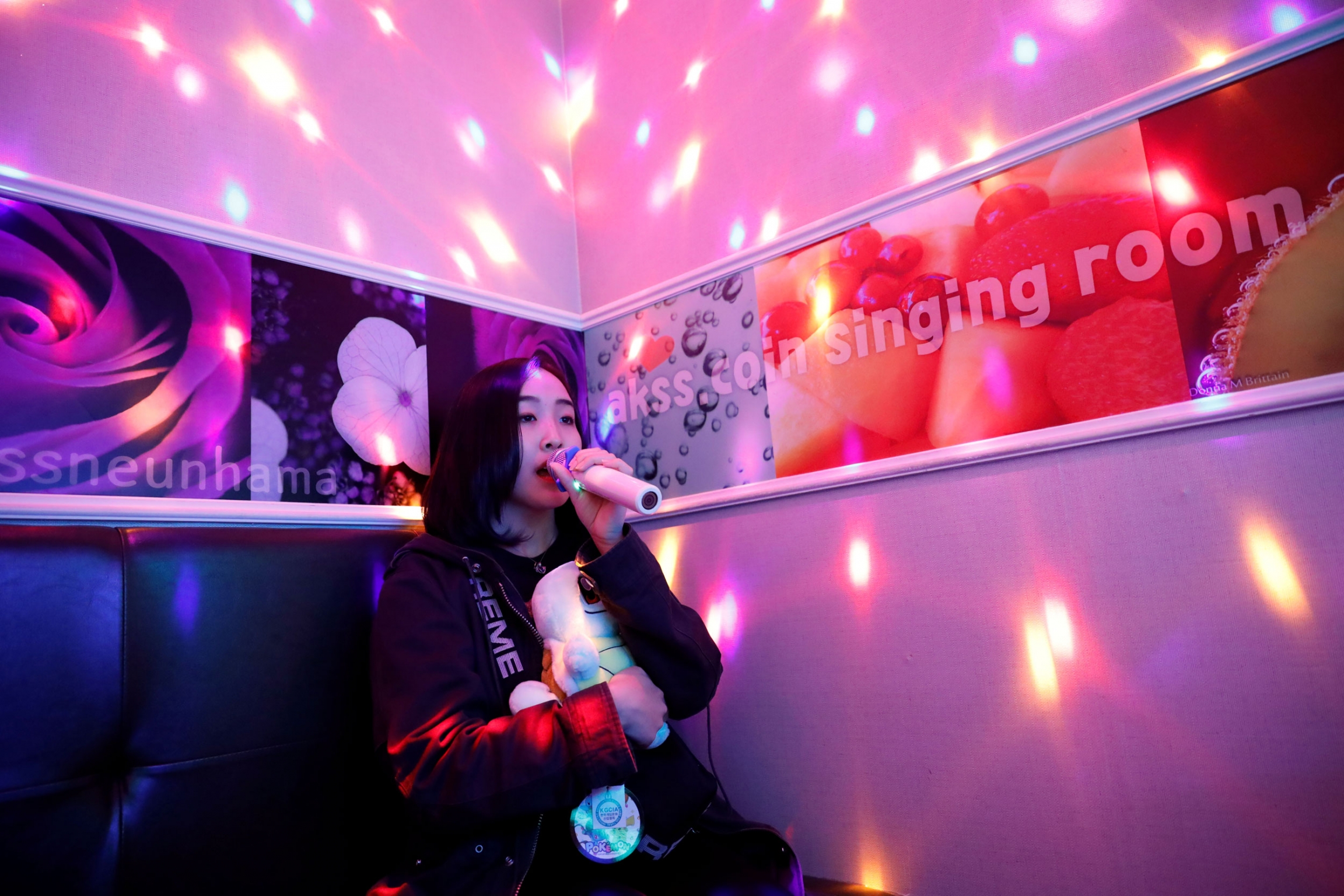 Hasumi sings as she spends time after class. A woman is shown singing into a microphone with pink lights on the walls.
