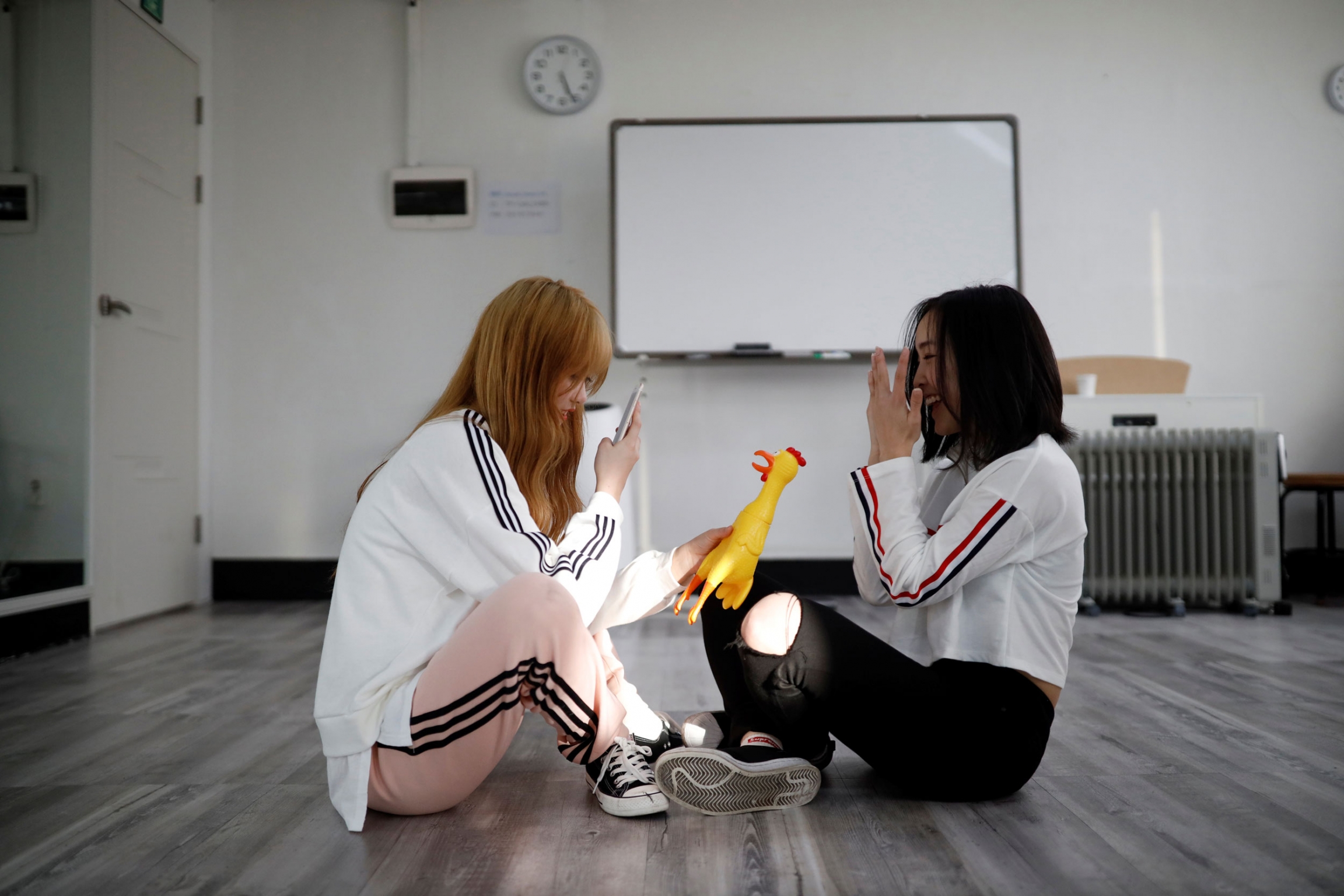 Wakamatsu takes photographs of Hasumi during a training session. Two young women are shown sitting on the floor with one holding a yellow rubber chicken and taking a photo with her mobile phone.