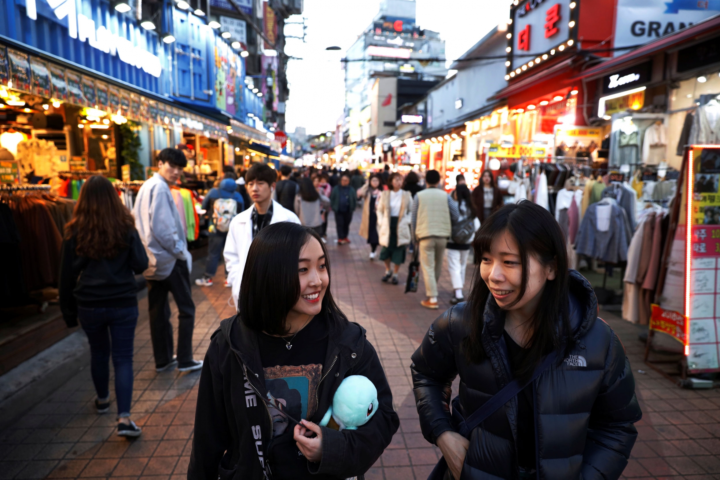 Hasumi walks with a manager of Acopia School. Two women are shown walking on a crowded street at dusk.
