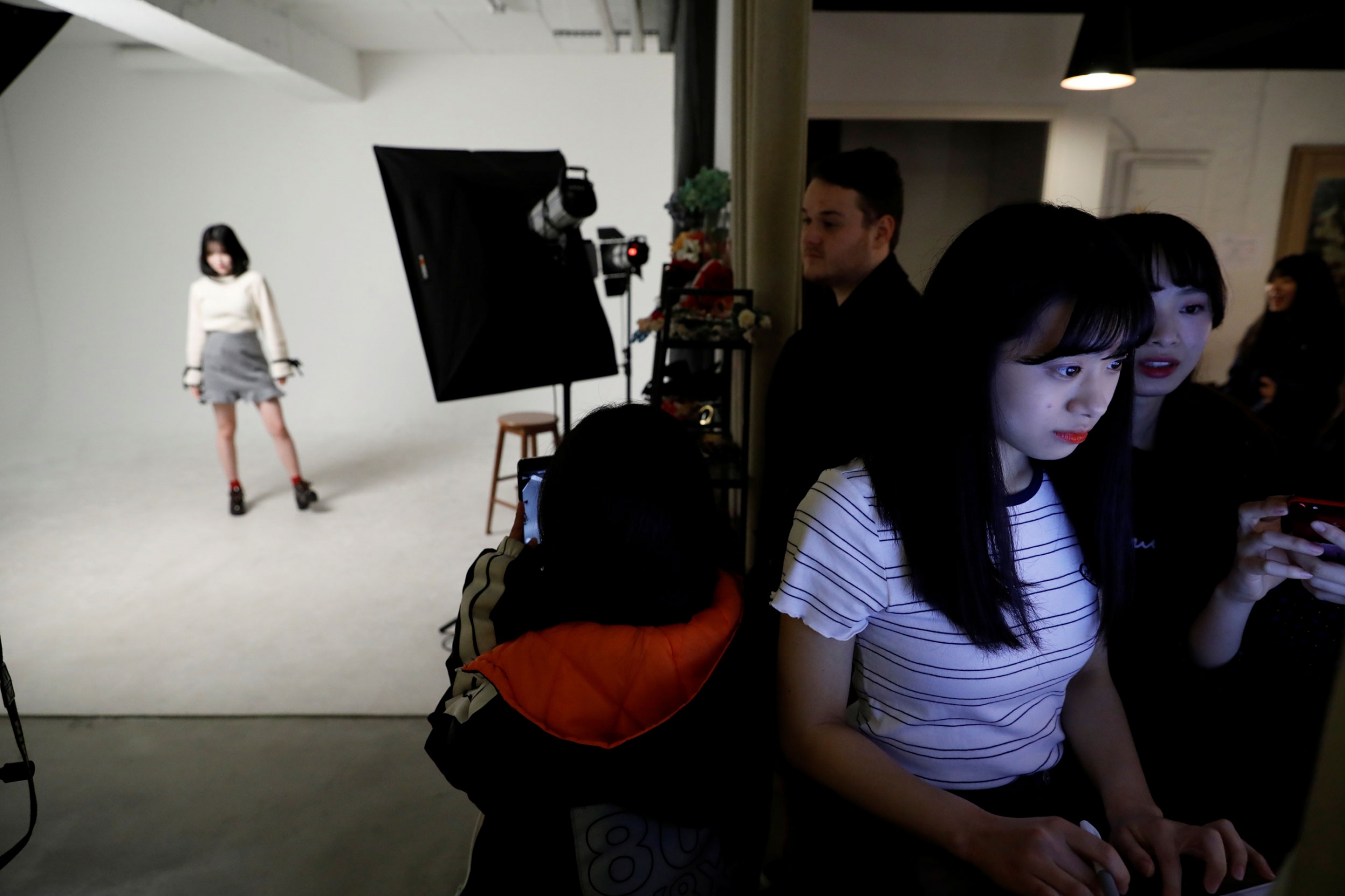 Niitsu chooses her profile picture before an audition. A woman is shown in the background posing for a photo with a white backgroup behind her and another woman in the near ground shown in the glow of a computer monitor.