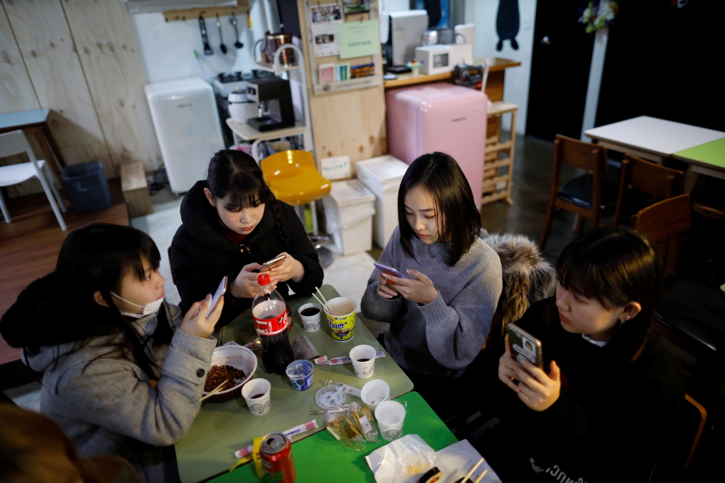 Yuuka Hasumi and her friends have lunch after a Korean language class in Seoul, South Korea. Four young women are shown sitting around two small green tables, all of them with a mobile phone.