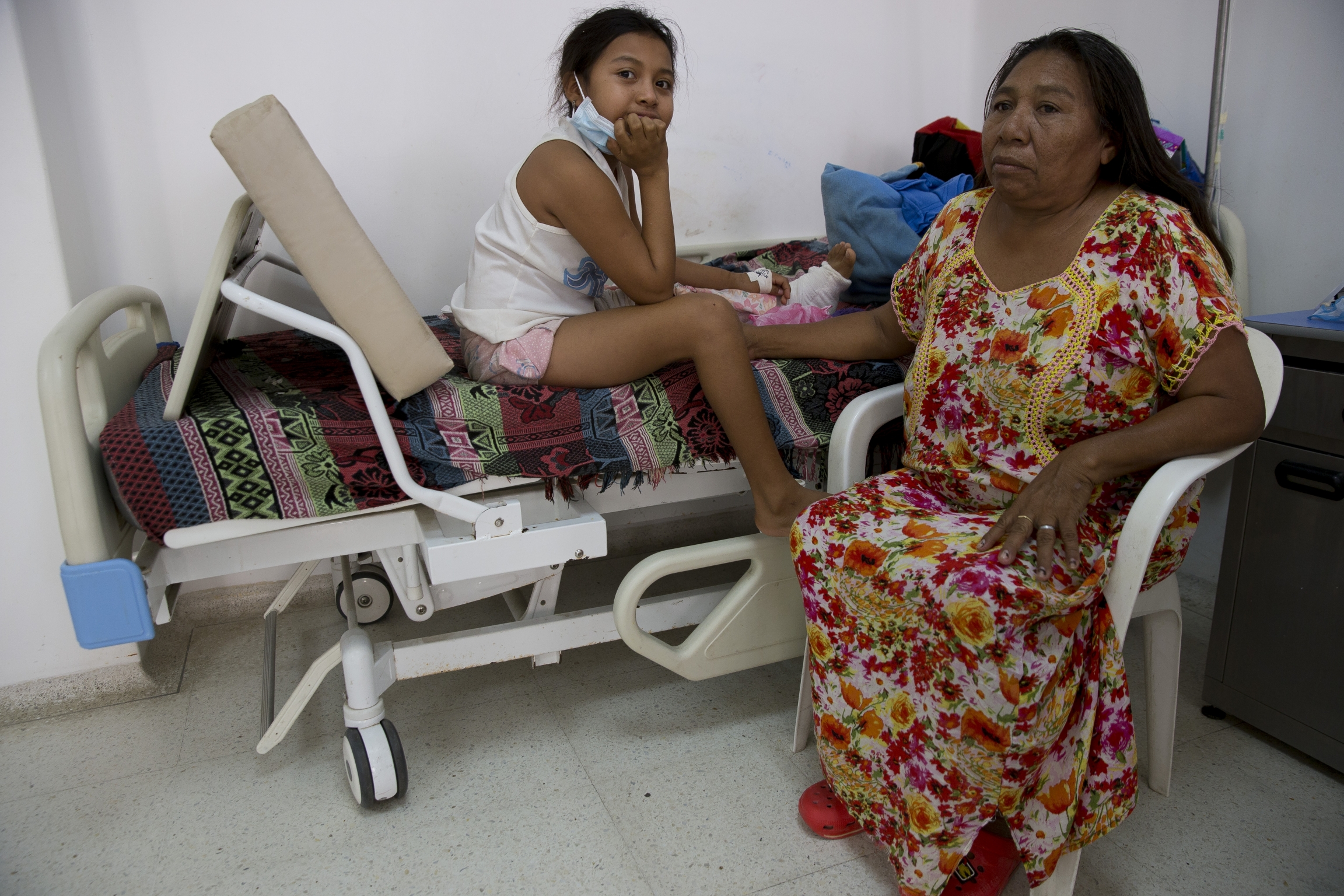 A young girl sits on a hospital bed with a fractured leg in bandages next to her mother.