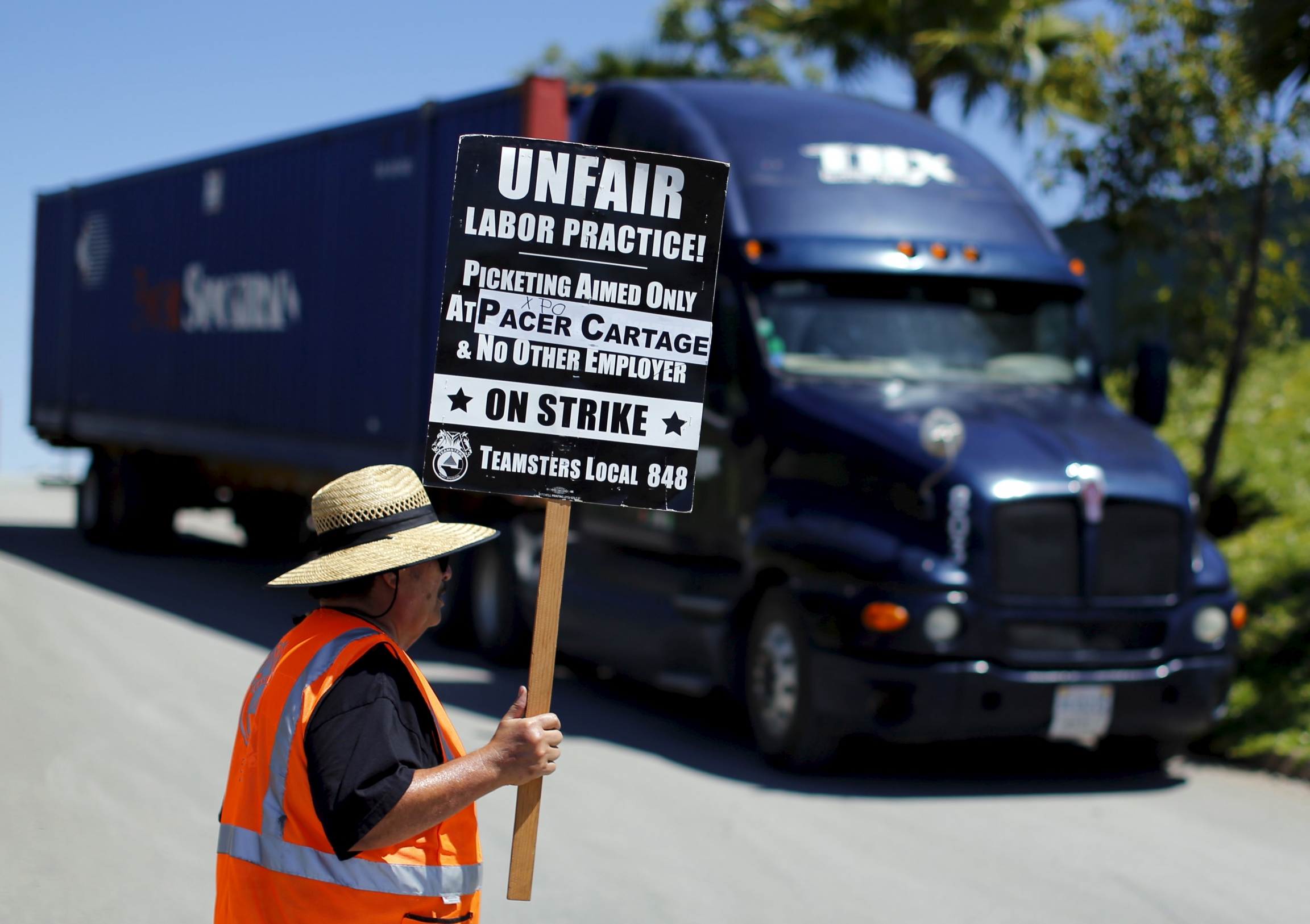 A striking truck driver walks a picket line at a Pacer Cartage facility in Otay Mesa, California. Drivers accused trucking companies of wage theft. A man next to a truck stands in an orange vest with a sign that says "unfair labor practice."