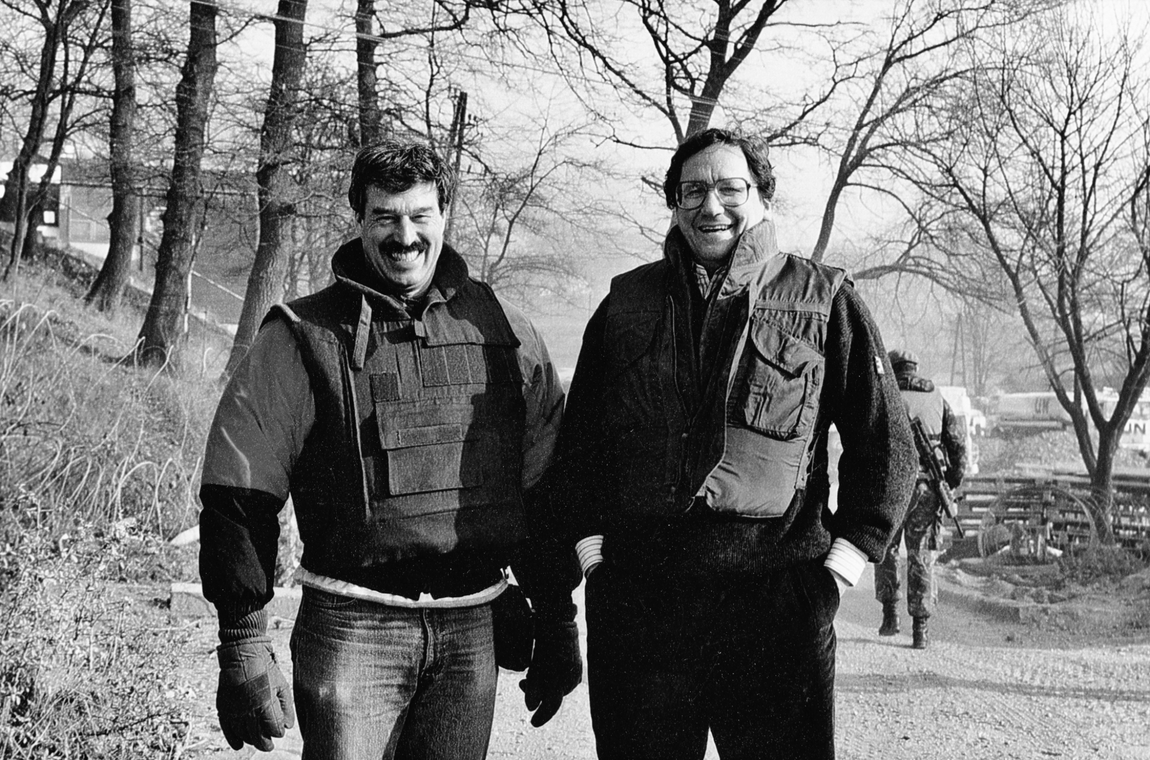 Richard Holbrooke, right, and Lionel Rosenblatt, then president of Refugees International, pose for a photo on the road to Sarajevo in the early 1990s. Two men stand in coats on a road.