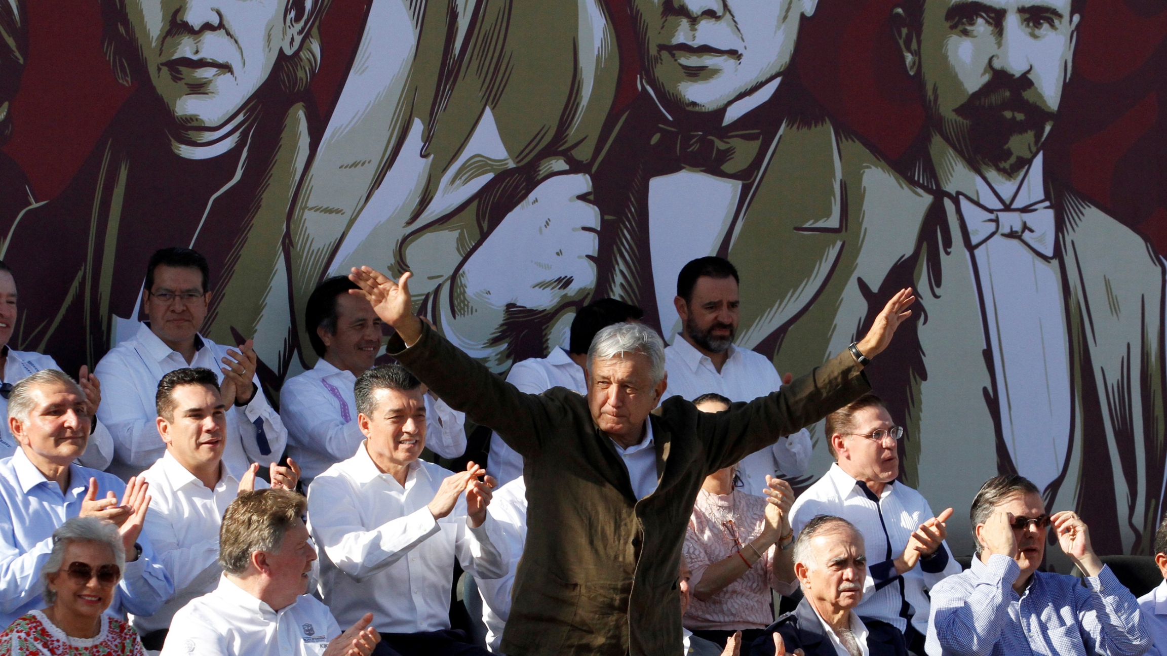Mexico's President Andrés Manuel López Obrador takes part on a "unity" rally to defend the dignity of Mexico and talks about the trade negotiations with the US in Tijuana, Mexico, on June 8, 2019. A man stands at a podium, his arms raised as he speaks. Others are seated around him.
