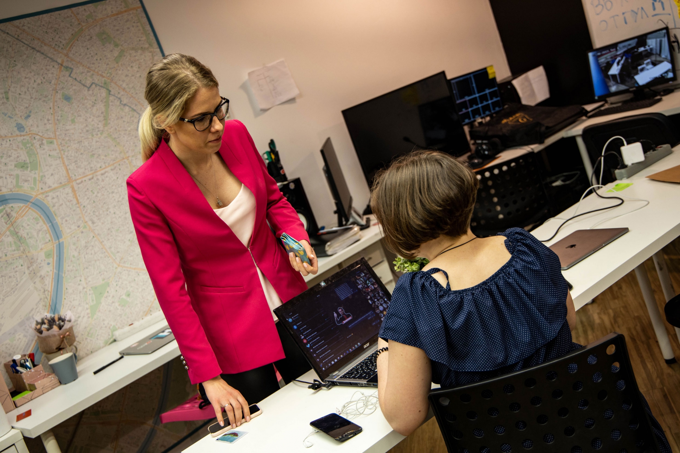 A woman in a pink blazer speaks with a colleague seated at a desk