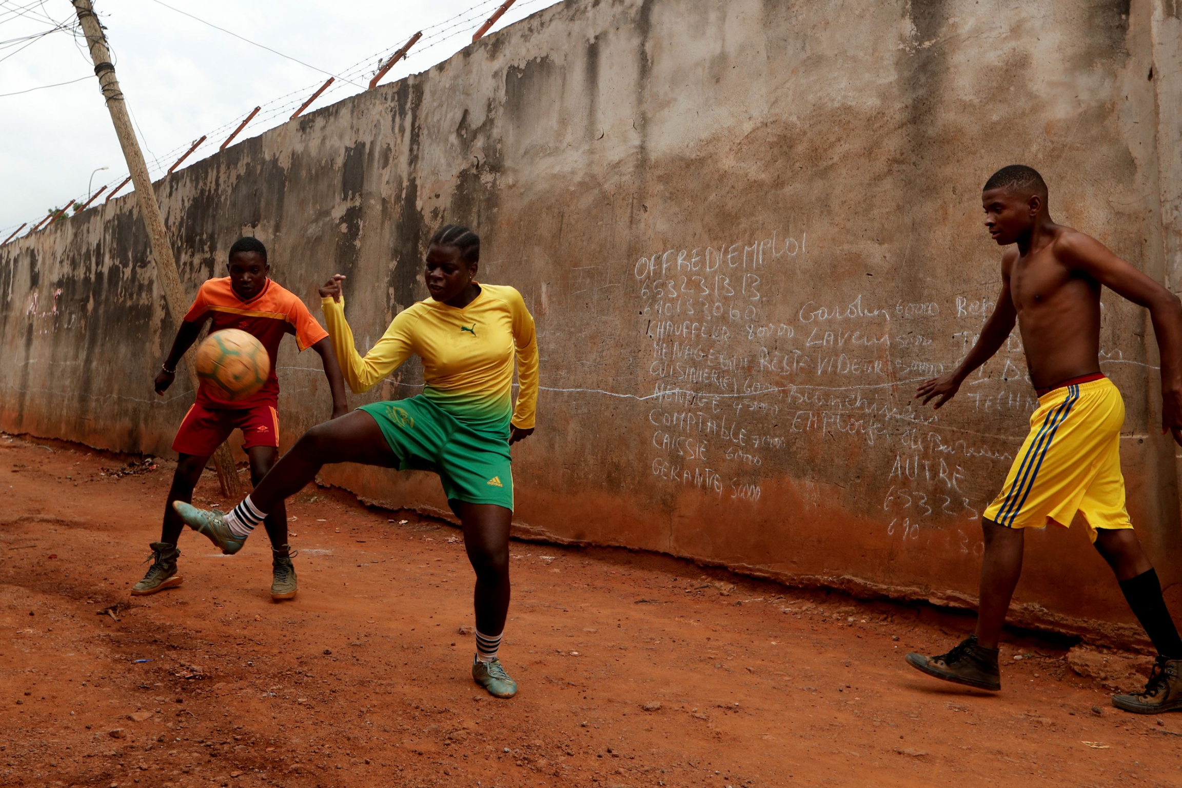 Asheri plays soccer with her friends outside her house. A young woman is shown juggling a soccer ball with two youn men.
