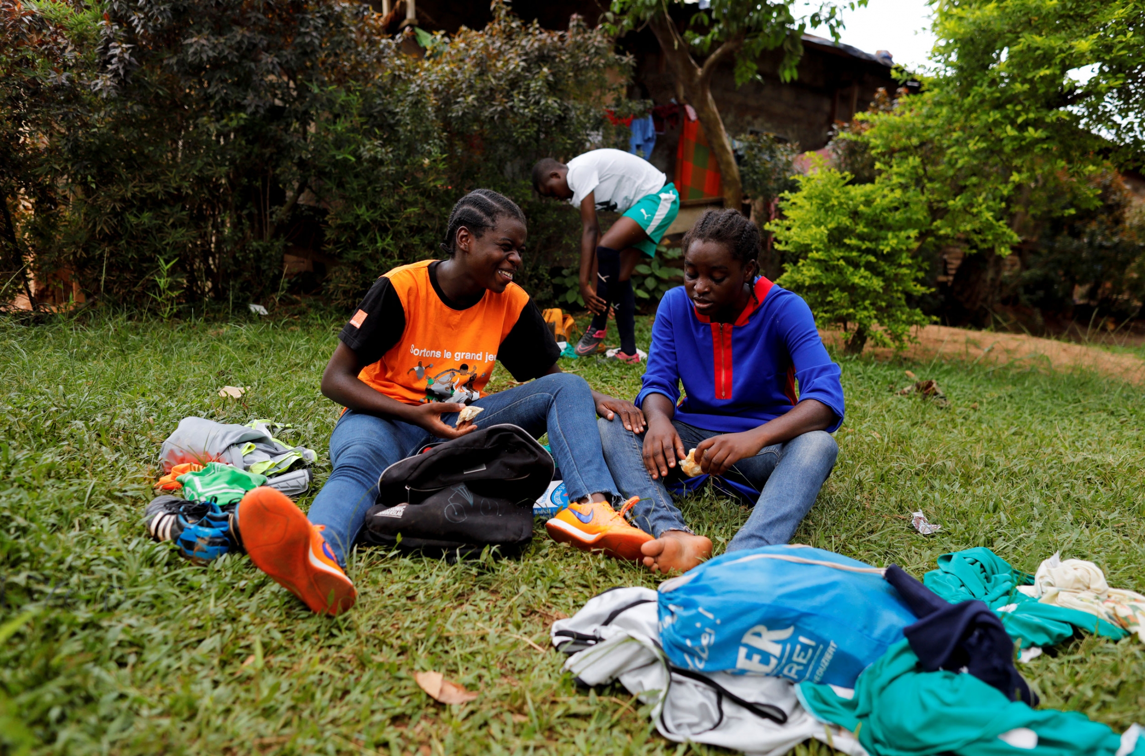 Asheri and Pouadjeu sit on the grass as they speak after their friendly match with male U15 team. Two young women are shown sitting in the grass smiling.