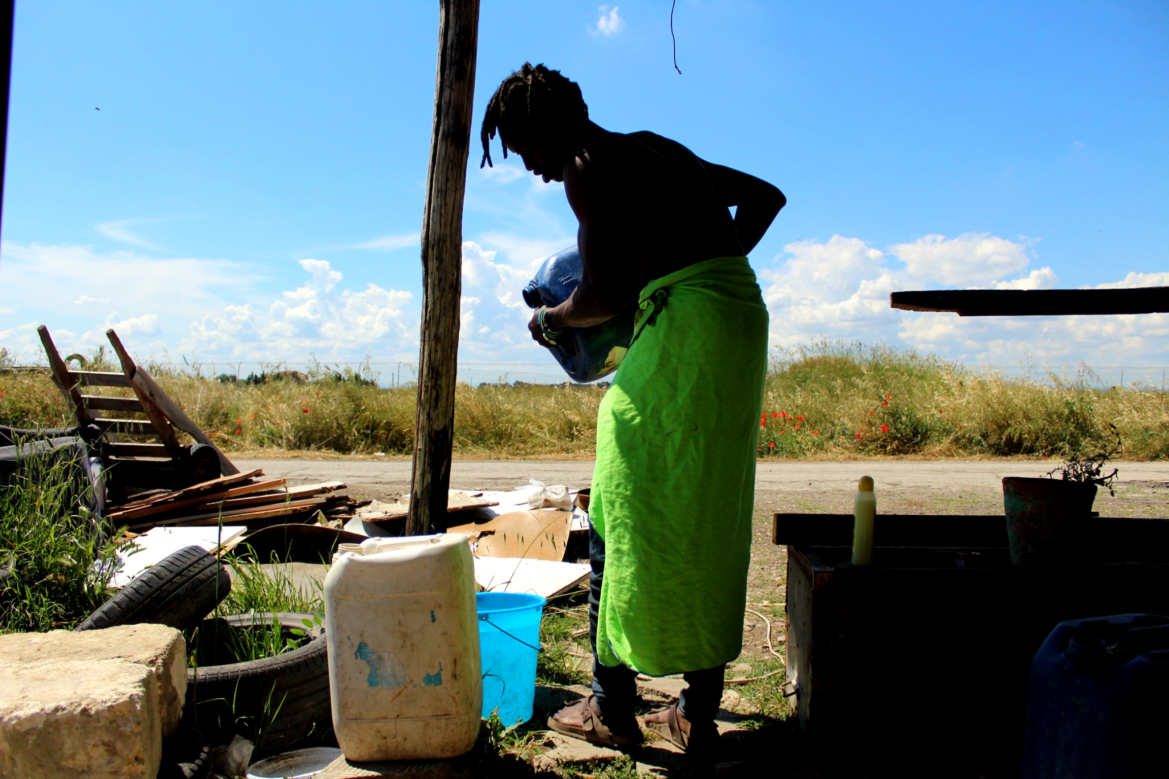 Young man uses jerry can to shower wearing green towel around waist. 