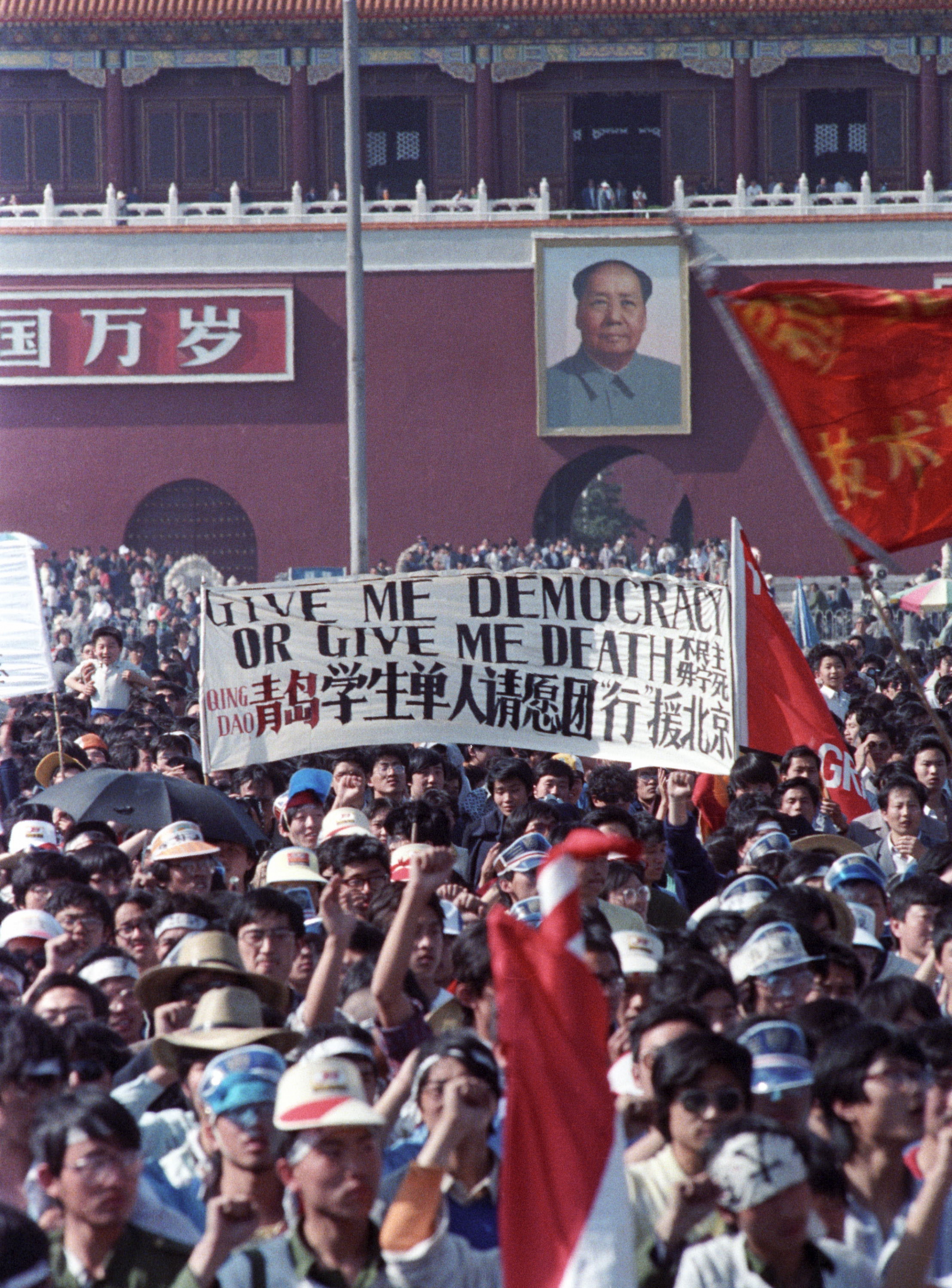 Chinese students carry a sign that reads "Give me democracy or give me death" during a demonstration in Tiananmen Square on May 14, 1989. The entrance to the Forbidden City and a portrait of Mao Zedong is in the background. Protesters crowd Tiananmen Square and carry a banner that says "Give me democracy or give me death."