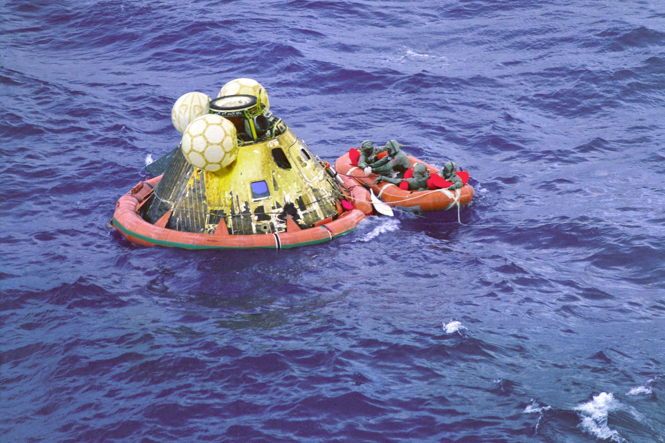 The Apollo 11 crew and a US Navy underwater demolition team swimmer await pickup by the USS Hornet recovery ship. A small orange raft is shown with astaurants aboard, floating next to luna modual.