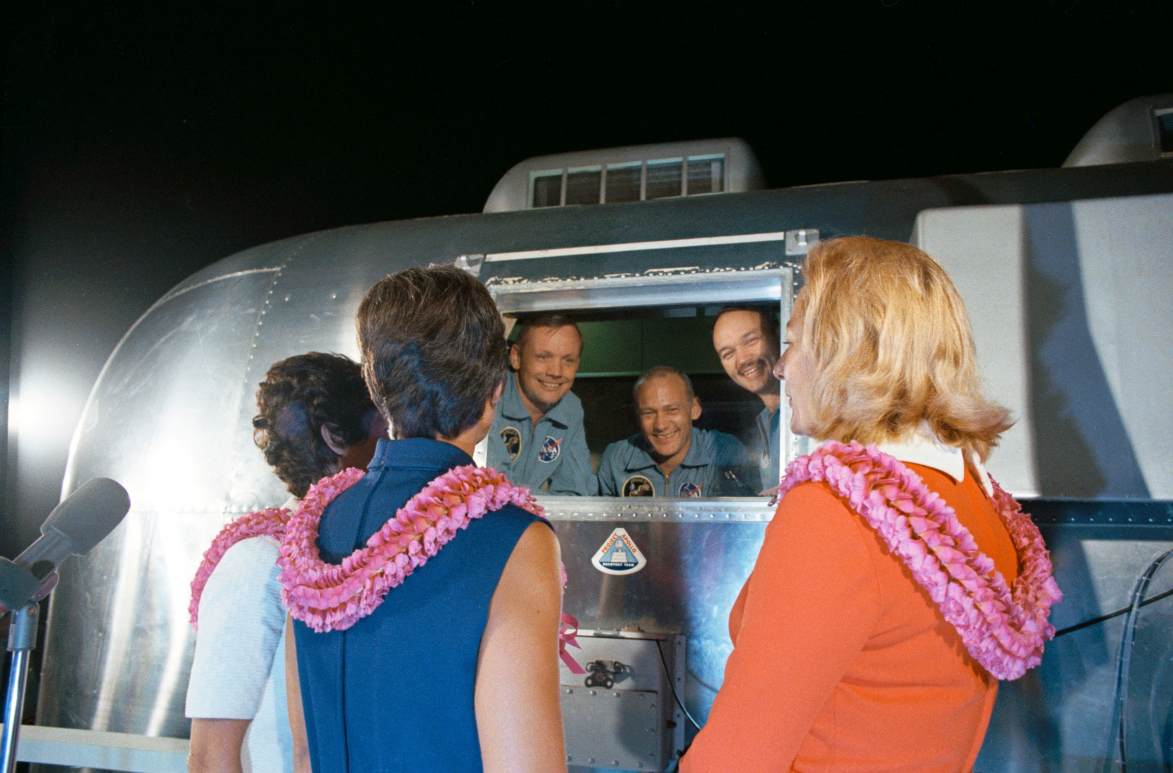 Looking through the window of a Mobile Quarantine Facility (MQF) are (L to R) astronauts Neil A. Armstrong, Edwin E. Aldrin Jr., and Michael Collins speak with their wives (L to R) Pat Collins, Jan Armstrong, and Joan Aldrin at Ellington Air Force Base, 1 A silver-colored containment structure is shown Neil A. Armstrong, Edwin E. Aldrin Jr., and Michael Collins speaking with their wives.