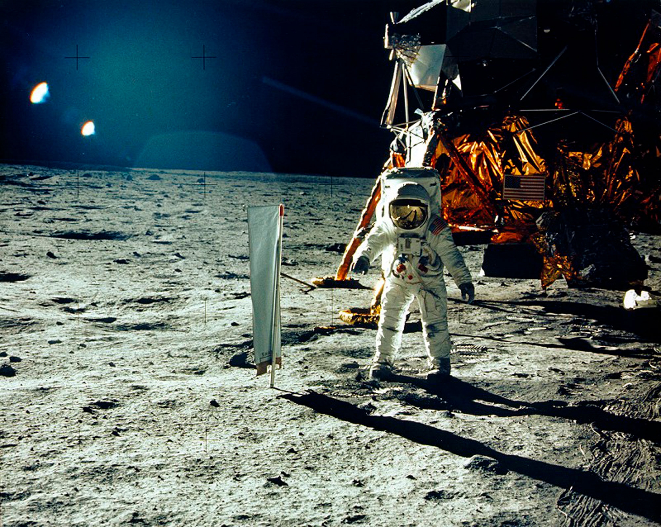 Buzz Aldrin stands beside a solar wind experiment next to the Lunar Module spacecraft on the surface of the Moon. An astraunant is show in full protective gear standing on the surface of the moon.