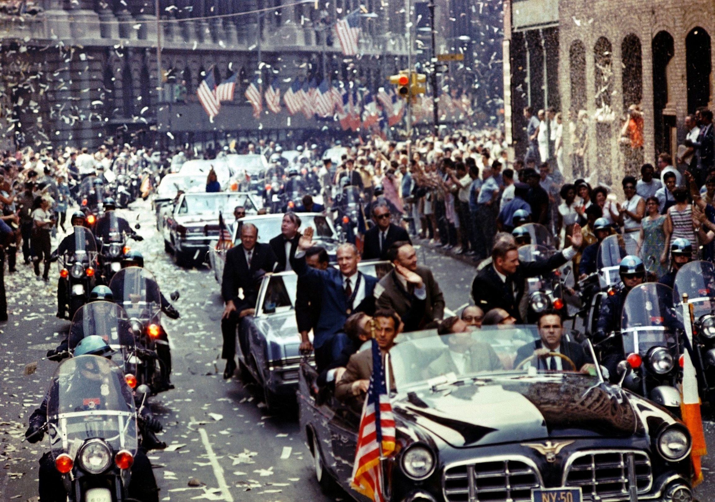 Apollo 11 lunar module pilot Buzz Aldrin, command module pilot Michael Collins and commander Neil Armstrong, seen in the lead car are showered in ticker tape during a parade down Broadway and Park Avenue in New York City, August 13, 1969. A row of cars is shown driving down the middle of the road with police motocycles on either side.
