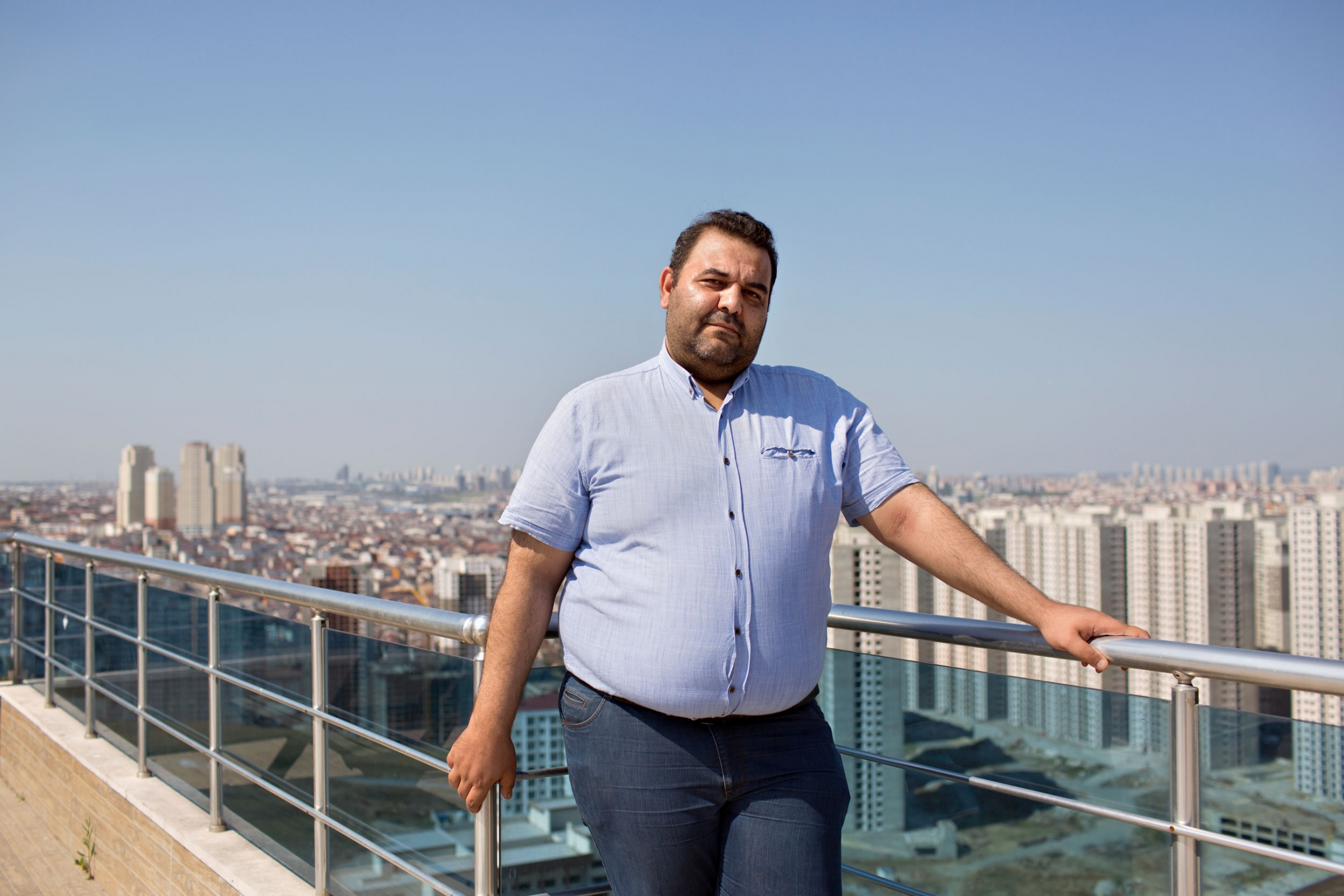 Mouath Youssef, an independent activist from Syria, is helping to track the arrests of Syrian refugees in Turkey. A man is shown in a blue button down shirt and jeans leaning against the railing of a rooftop deck.