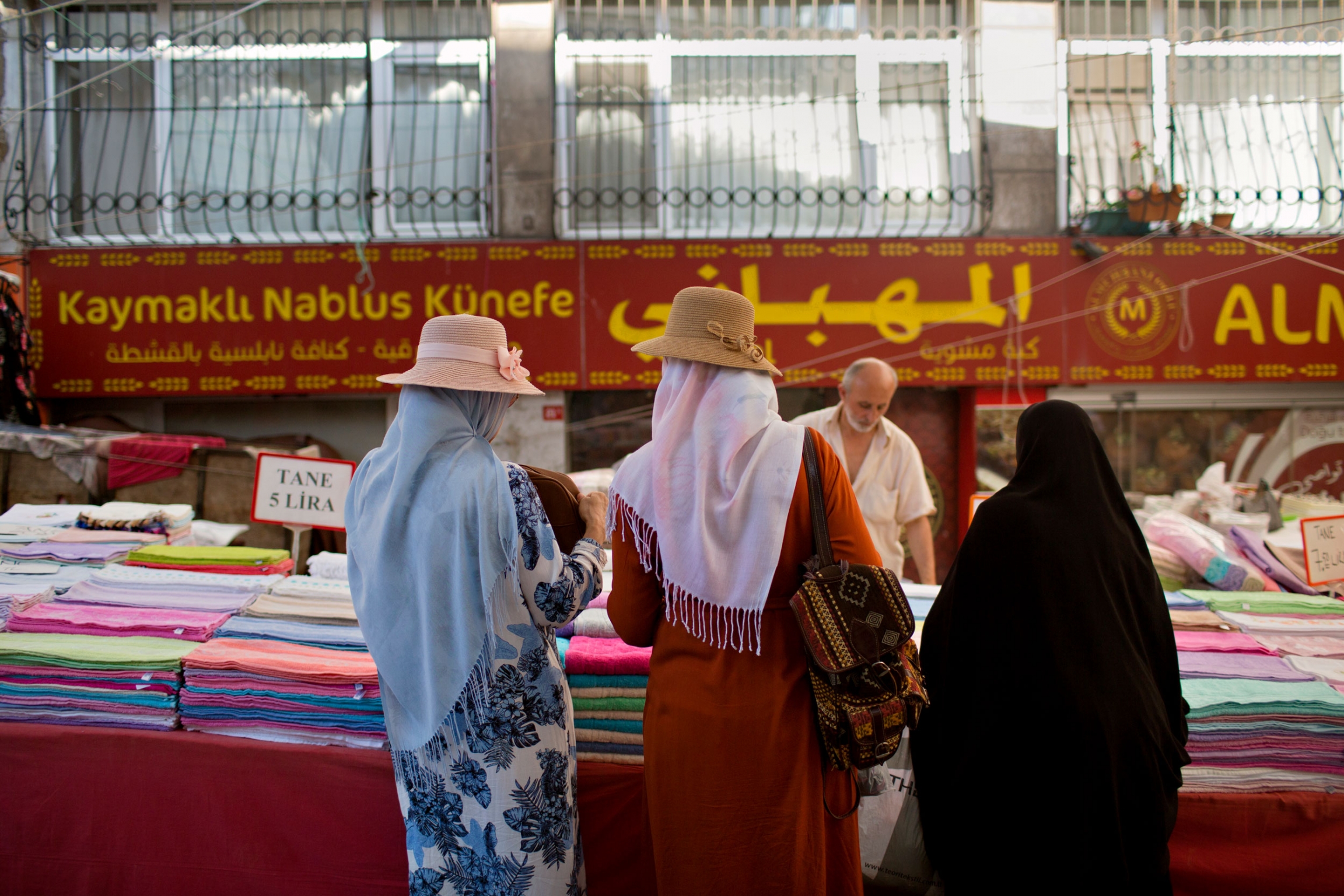 People shop in the Malta Market in the Fatih neighborhood of Istanbul, which has a large Syrian population. Three women are shown standing at a table with scarves.