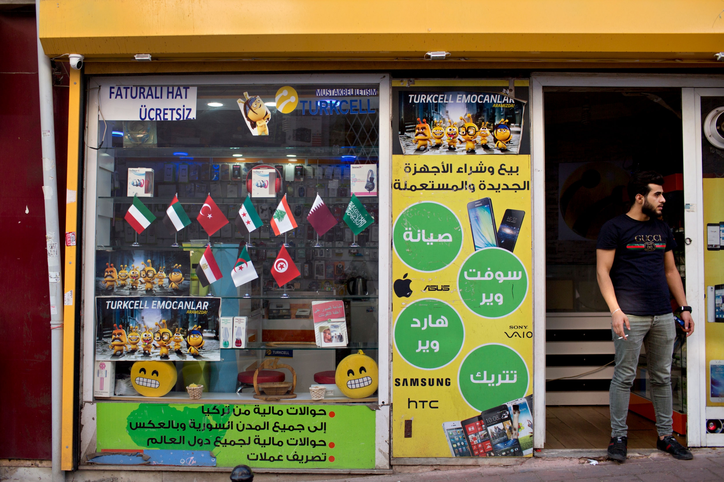 A man smokes a cigarette in front of a store with words written in Arabic in the Aksaray neighborhood of Istanbul, which has a large Syrian population. A man in a black t-shirt is shown standing in the doorway of a shop smoking a cigarette.