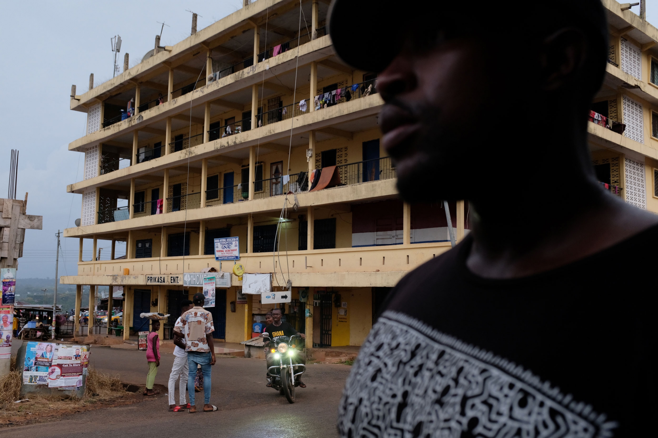 A man walks past a torn-down colonial building in Mampong, Ashanti Region, Ghana. A man is shown in the nearground looking off with a six story building with balconies in the background.
