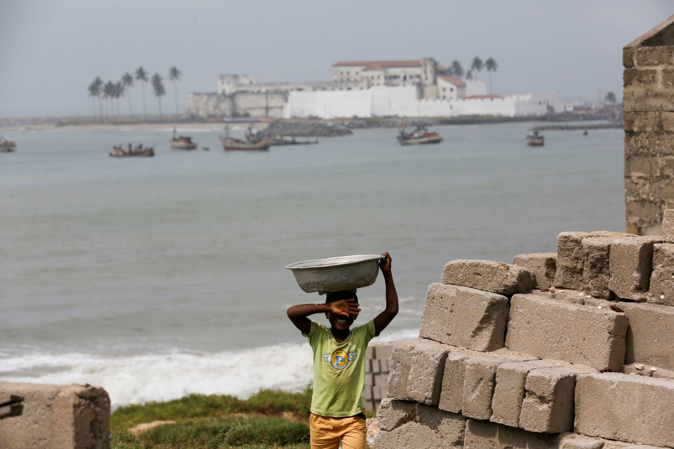 Elmina Castle is seen in the background. A young boy is shown in the nearground holding a large pan on his head with the ocean and Elmin Castle in the background.