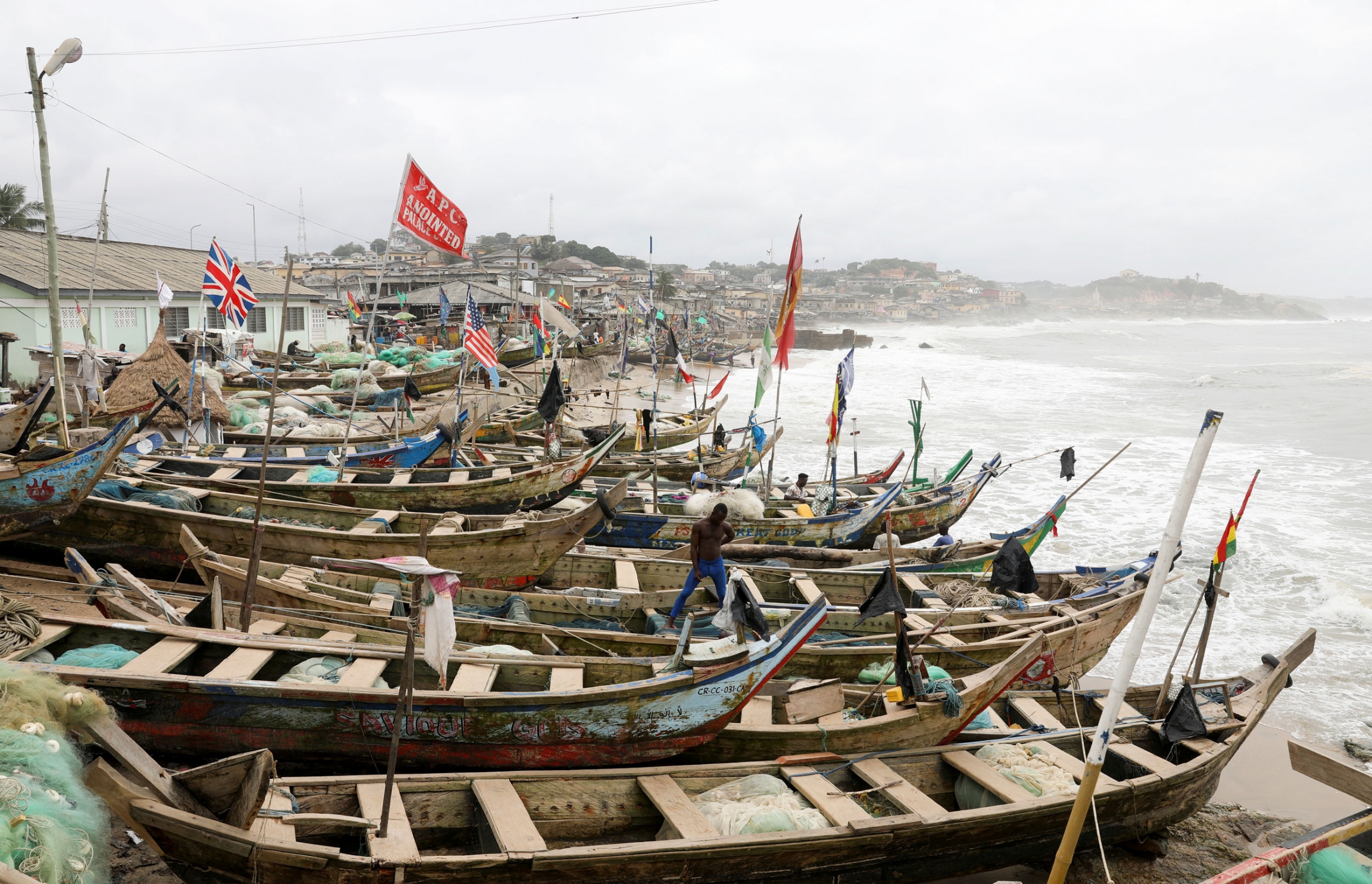 British and American flags are among other flags hoisted on fishermen's boats at the Cape Coast Castle. Several long wooden boats are shown in a row on the beach.