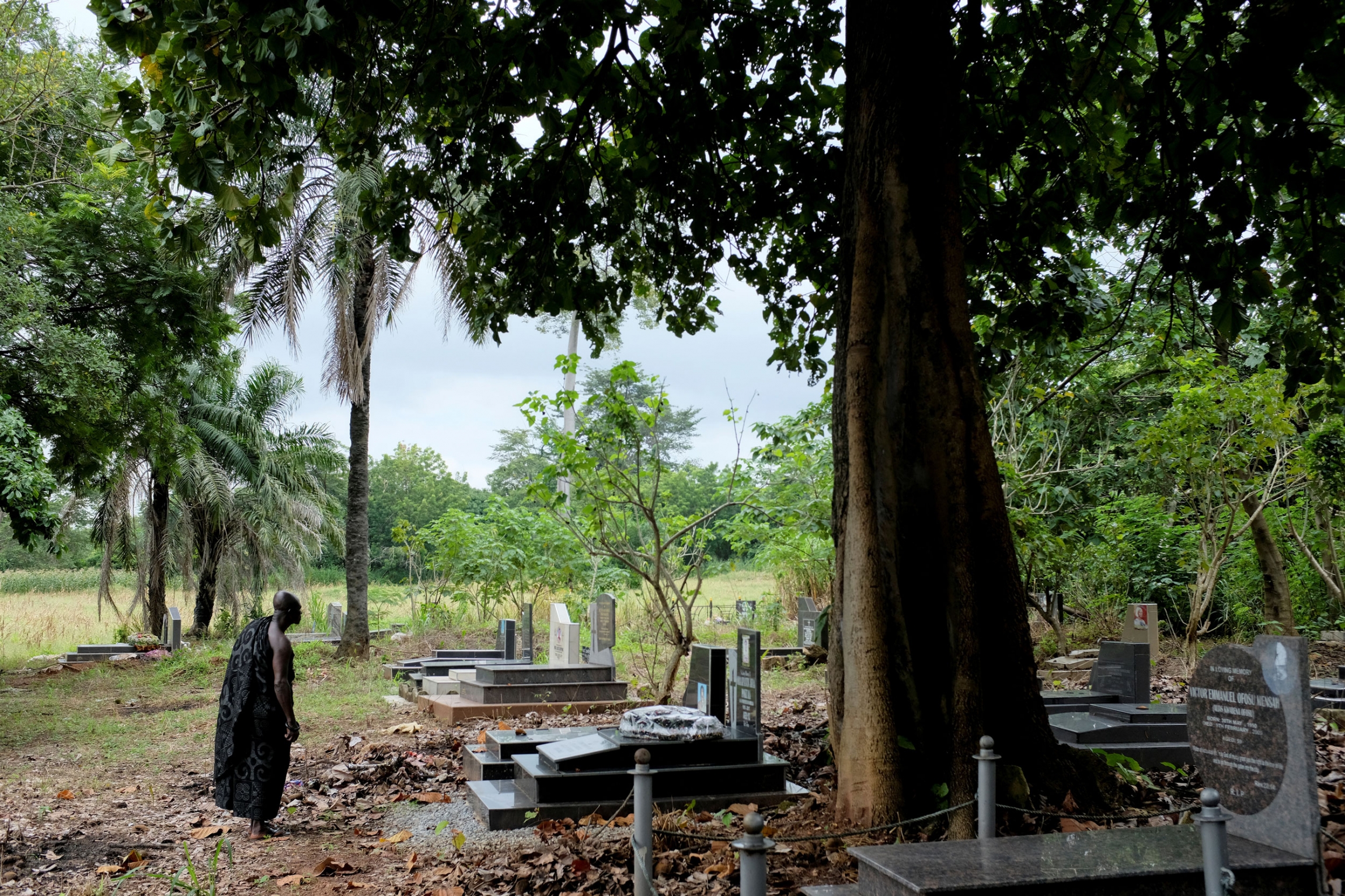 Assenso visits the grave of his uncle. A grave yard is shown with a man standing looking at a long, rectangular marble tombstone.