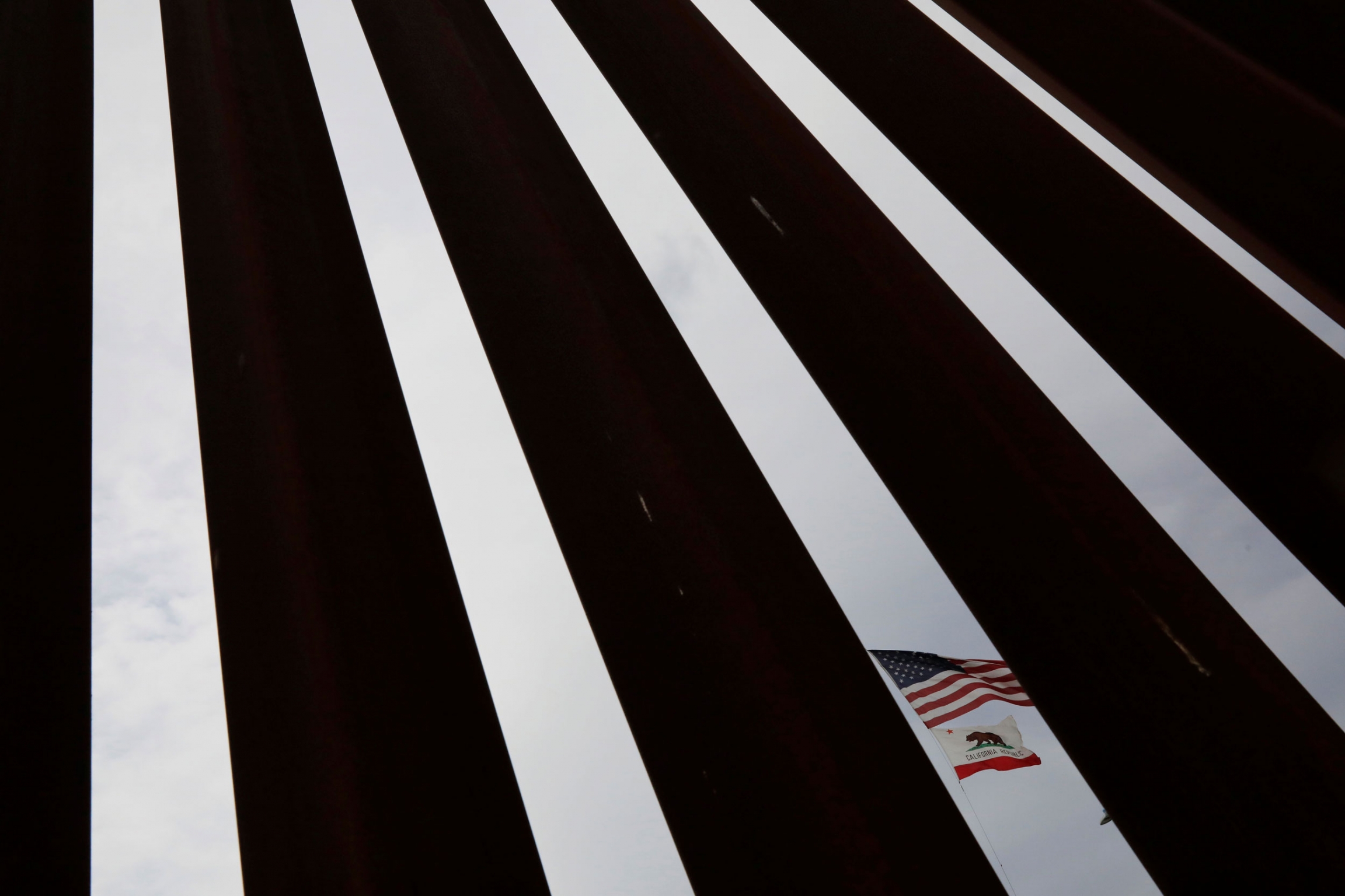 A US flag is seen through the border fence between Mexico and US in Tijuana, Mexico. A photograph with dark metal slats are shown in contrast to the blue sky in-between with the US and California flags in one of the slots.
