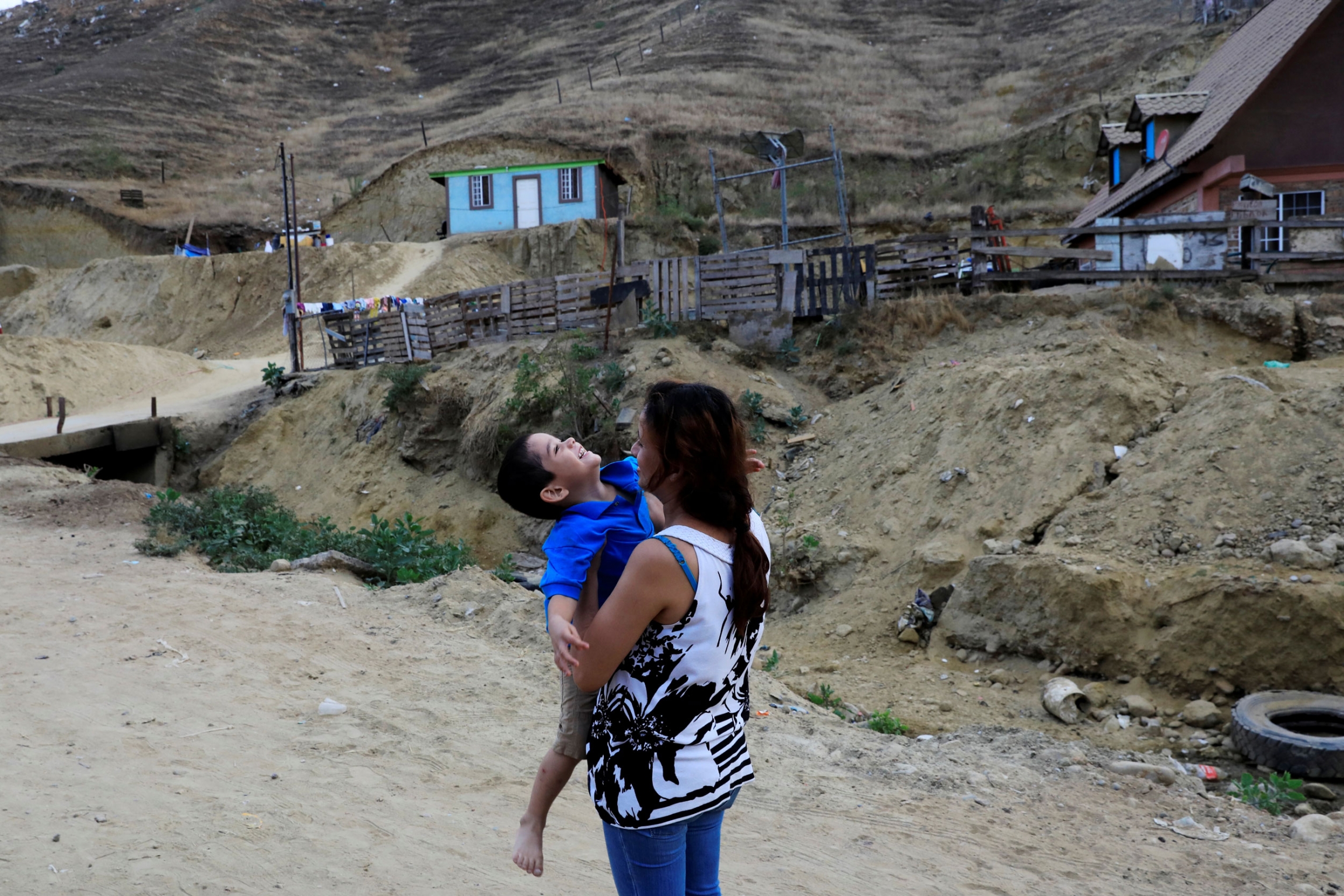 Migrant from Honduras, Dunia Ayala, carries one of her children outside a shelter after she was sent by the Customs and Border Protection to Mexico from the US under the MPP. A woman is show holding her small son who is leaning back and smiling.
