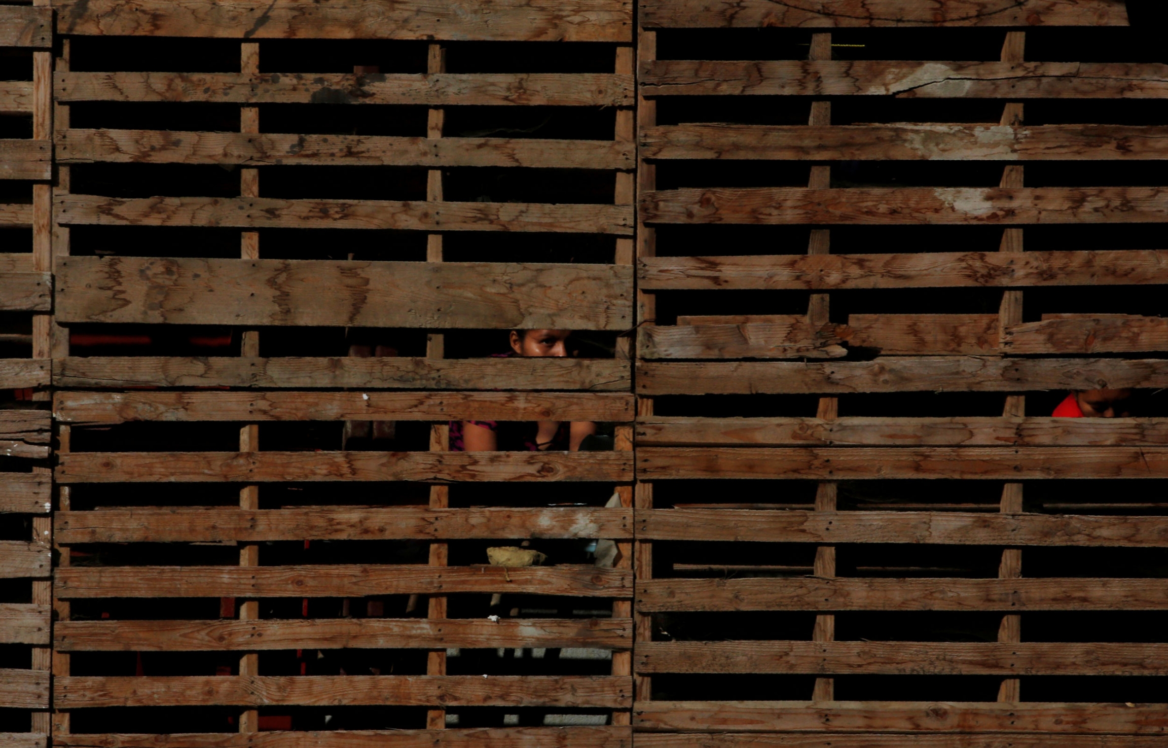 A migrant woman washes her clothes inside a shelter. Horizontal wooden slats are shown across the entire photograph with the face of a woman in shadown.