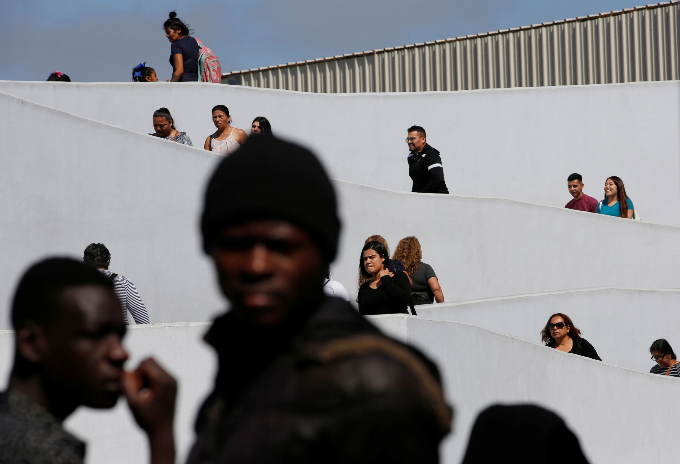 People cross the borders as migrants from different countries wait to apply for asylum in the US. Dozens of people are shown at various heights along a ramp zig-zagging up.