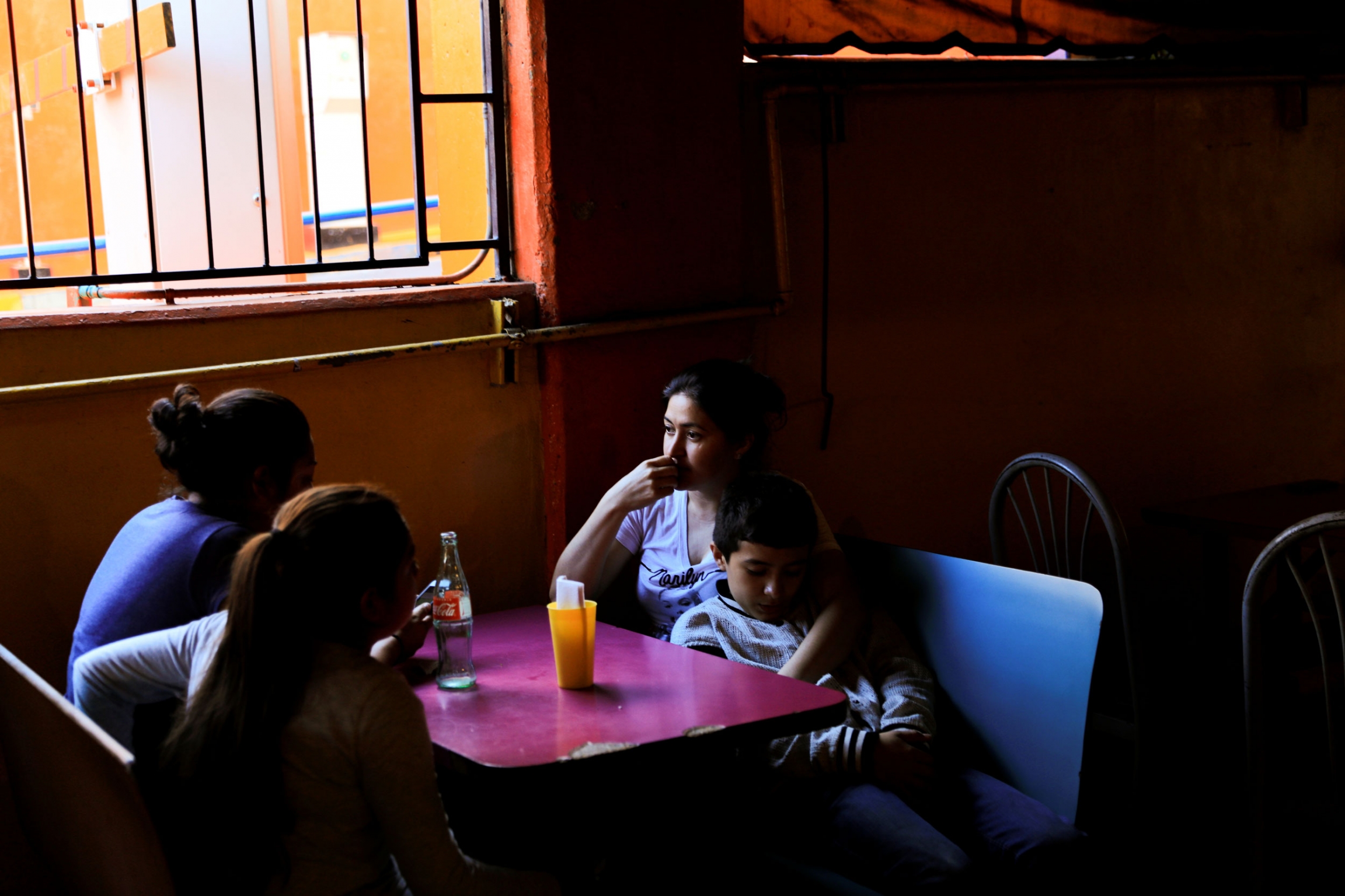 Migrant from Honduras, Patricia Acosta, 36, embraces her son Adalberto Acosta, 13, as they finish eating at a restaurant after they were sent by the Customs and Border Protection to Mexico from the US under the Migrant Protection Protocols (MPP), in Tijua Three women are shown sitting around a table along with a teenage boy shown by the light of a window from above.