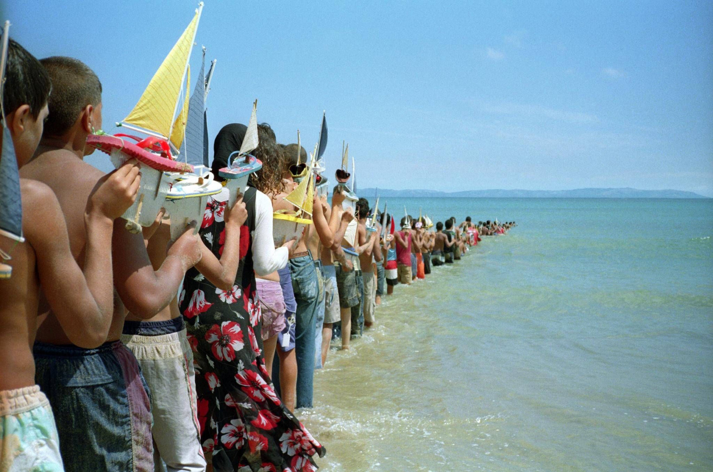 A still from the video “Don’t Cross the Bridge Before You Get to the River (Strait of Gibraltar, Morocco- Spain)." A line of people holding toy boats is seen going into the ocean.