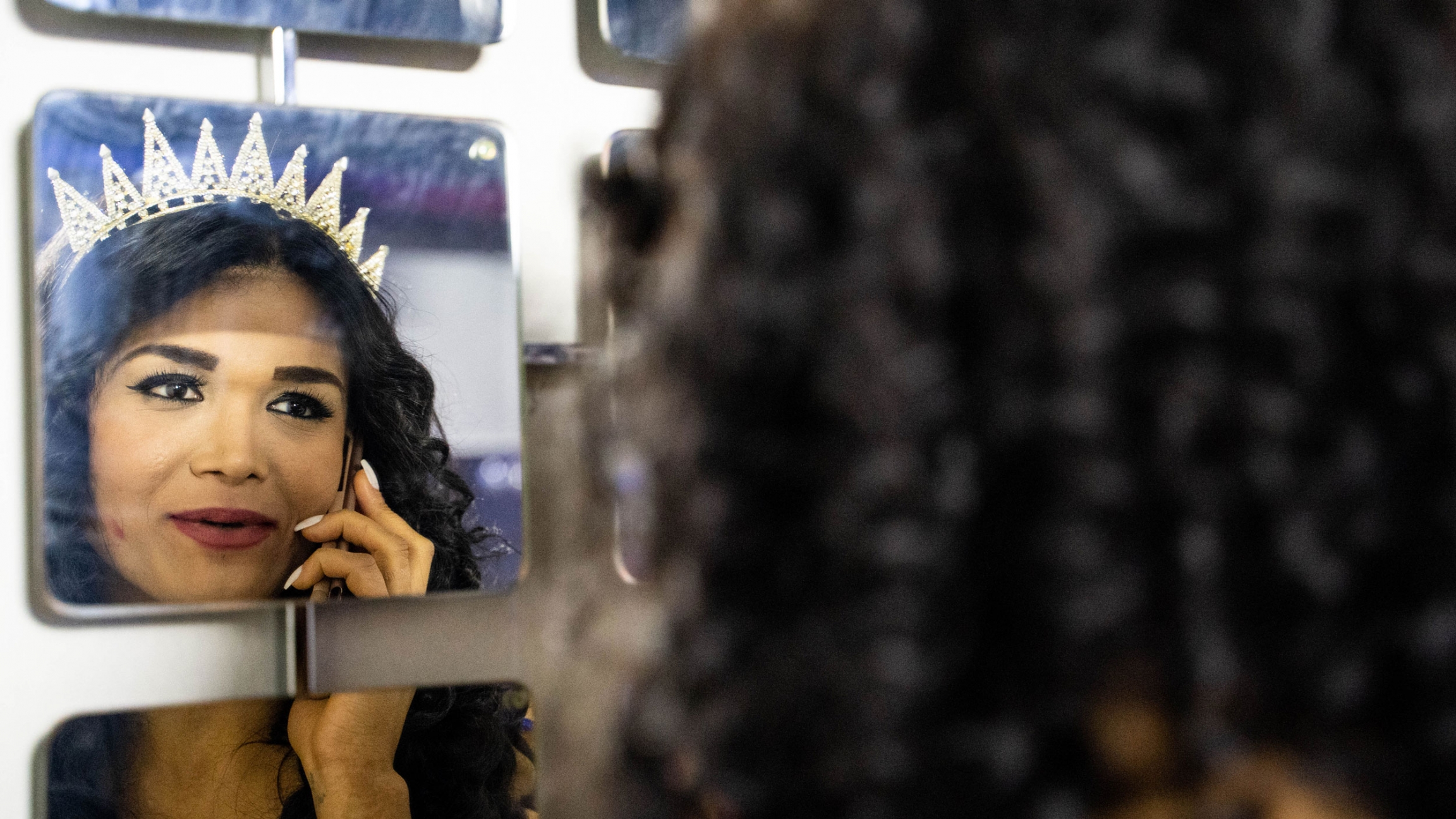 Didem Akay, on the phone with her family, gives them the news that she won the pageant. A transgender woman is shown in the reflection of a mirror holding a phone to her hear and wearing a crown.