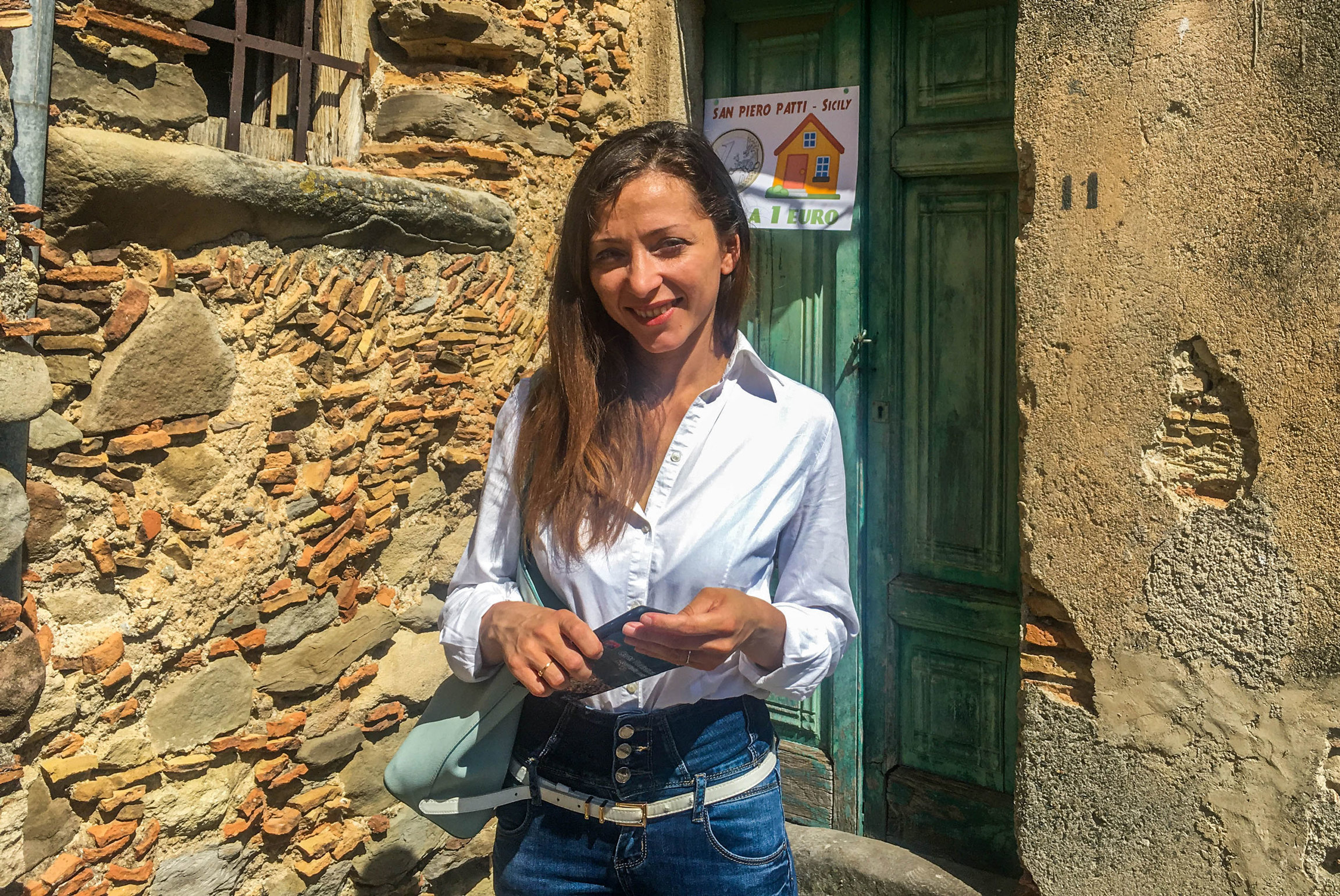 Katia Nepumaceno plans to make San Piero Patti, a small town in Italy, her permanent home. A woman is shown wearing a white button-down shirt and standing in front of a green door.