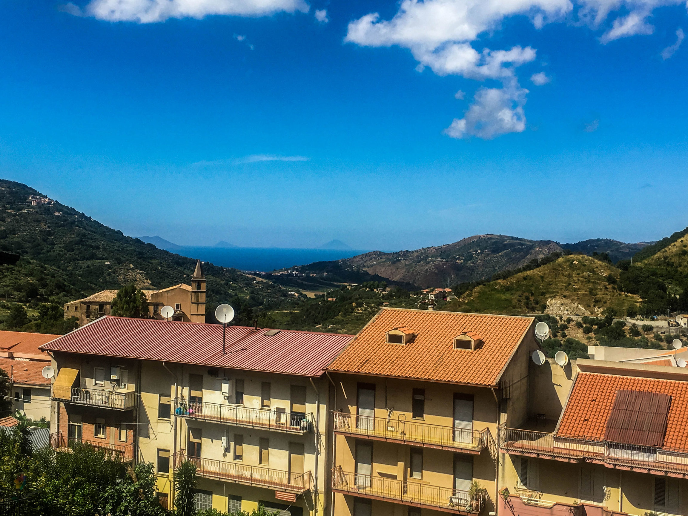 San Piero Patti, a small Italian town, is taking extreme measures to try and bring new life to the region — including selling some of its abandoned houses for less than a cup of coffee. Several old Italian houses are shown in a row with a forested hillside in the background.